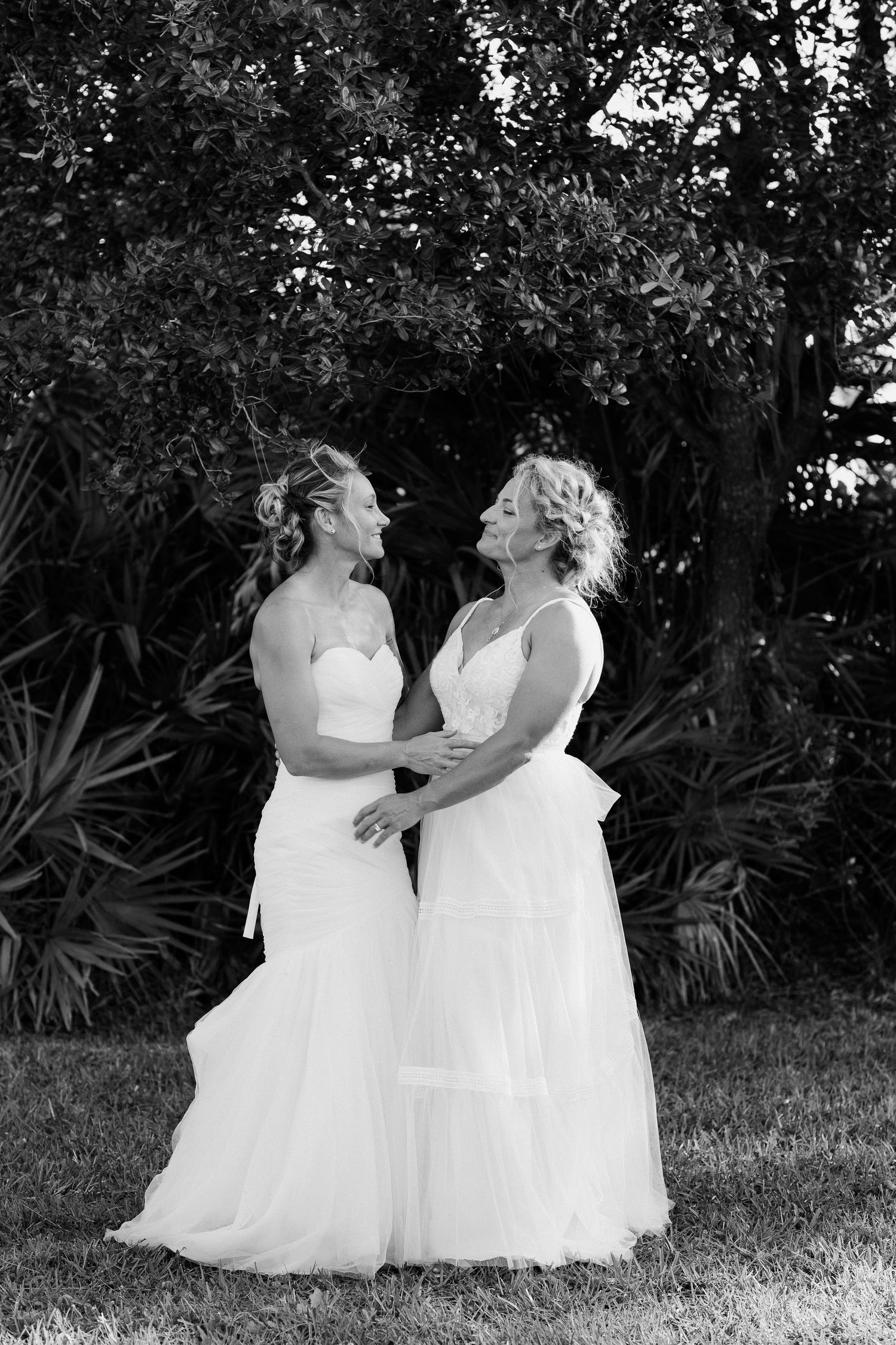 Two brides in white gowns embrace and laugh together in a black and white outdoor wedding portrait.