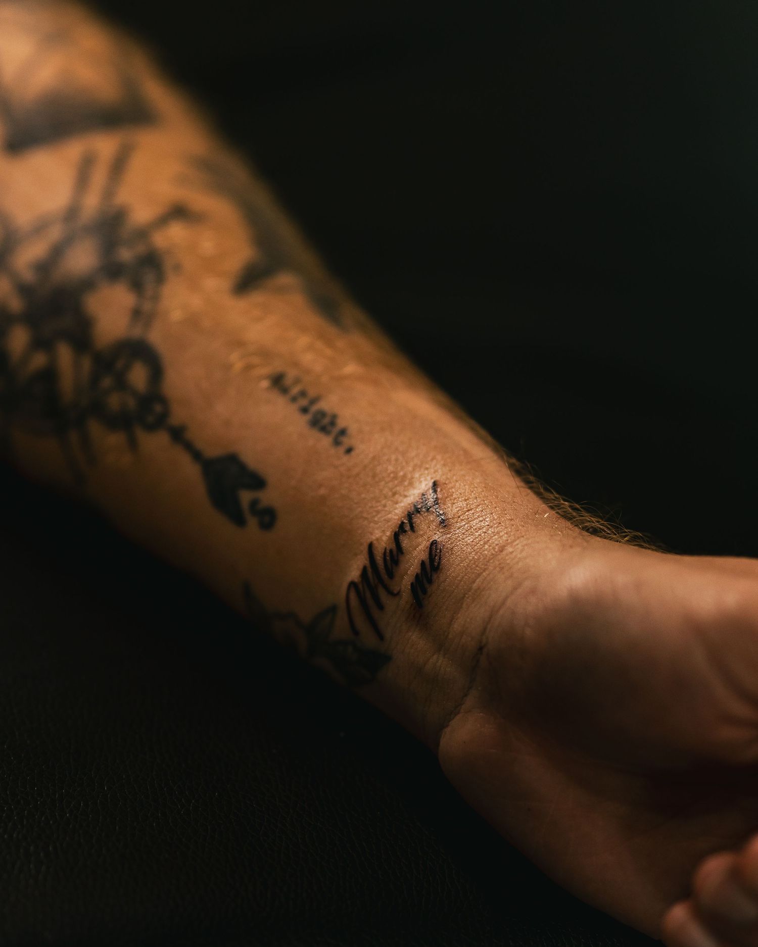 Close-up of a tattooed forearm with script and decorative ink designs on dark background.