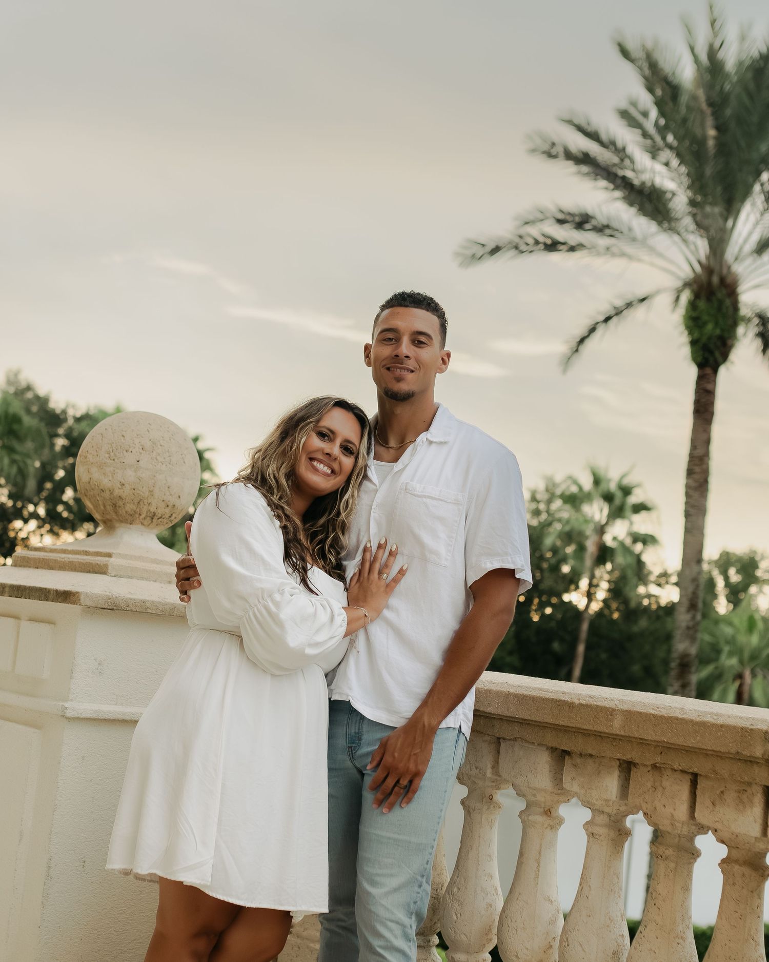Smiling couple in white outfits embracing on a balcony with palm trees at sunset.