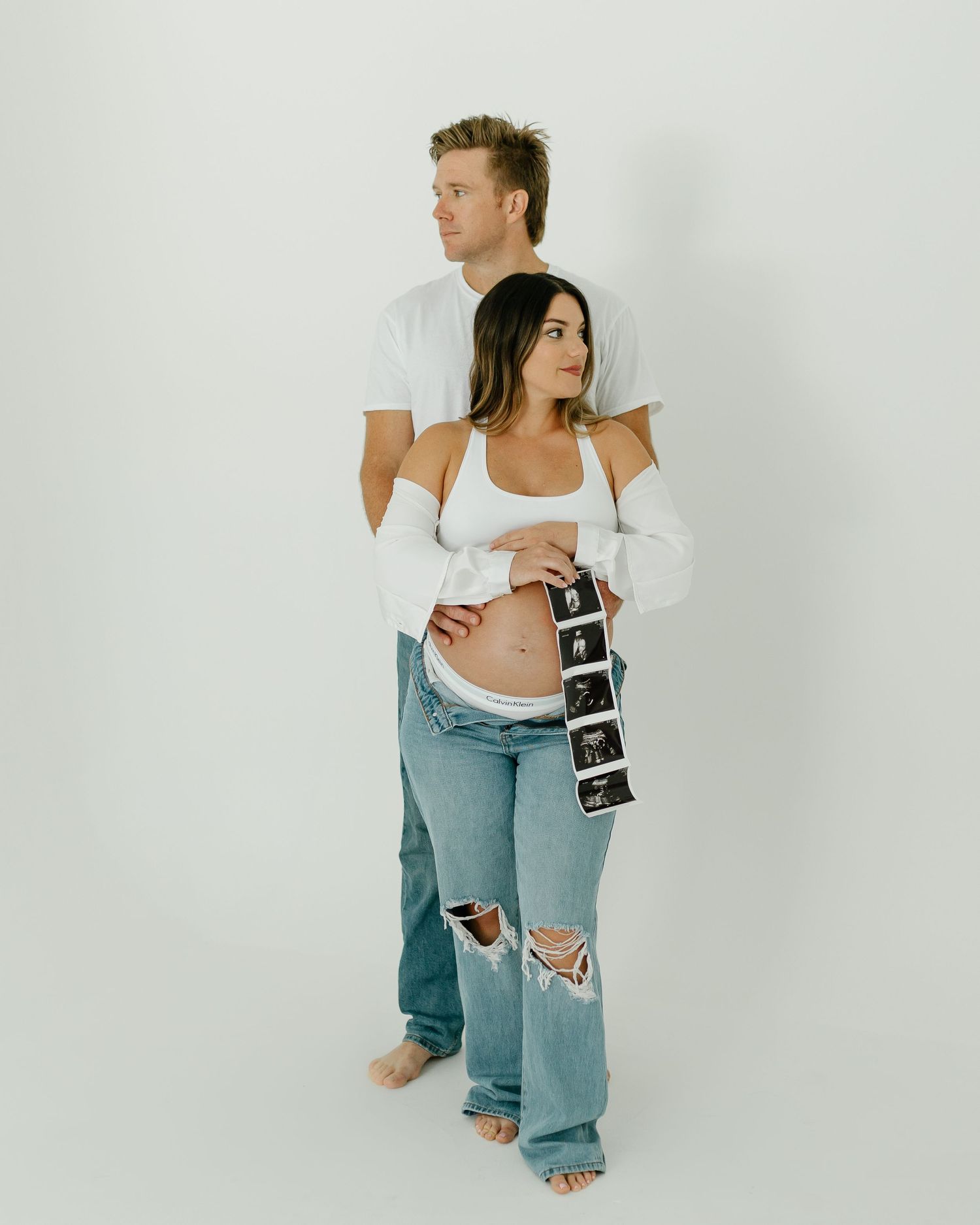 Expectant couple in white tops and jeans holding ultrasound photos during a bright studio maternity shoot.