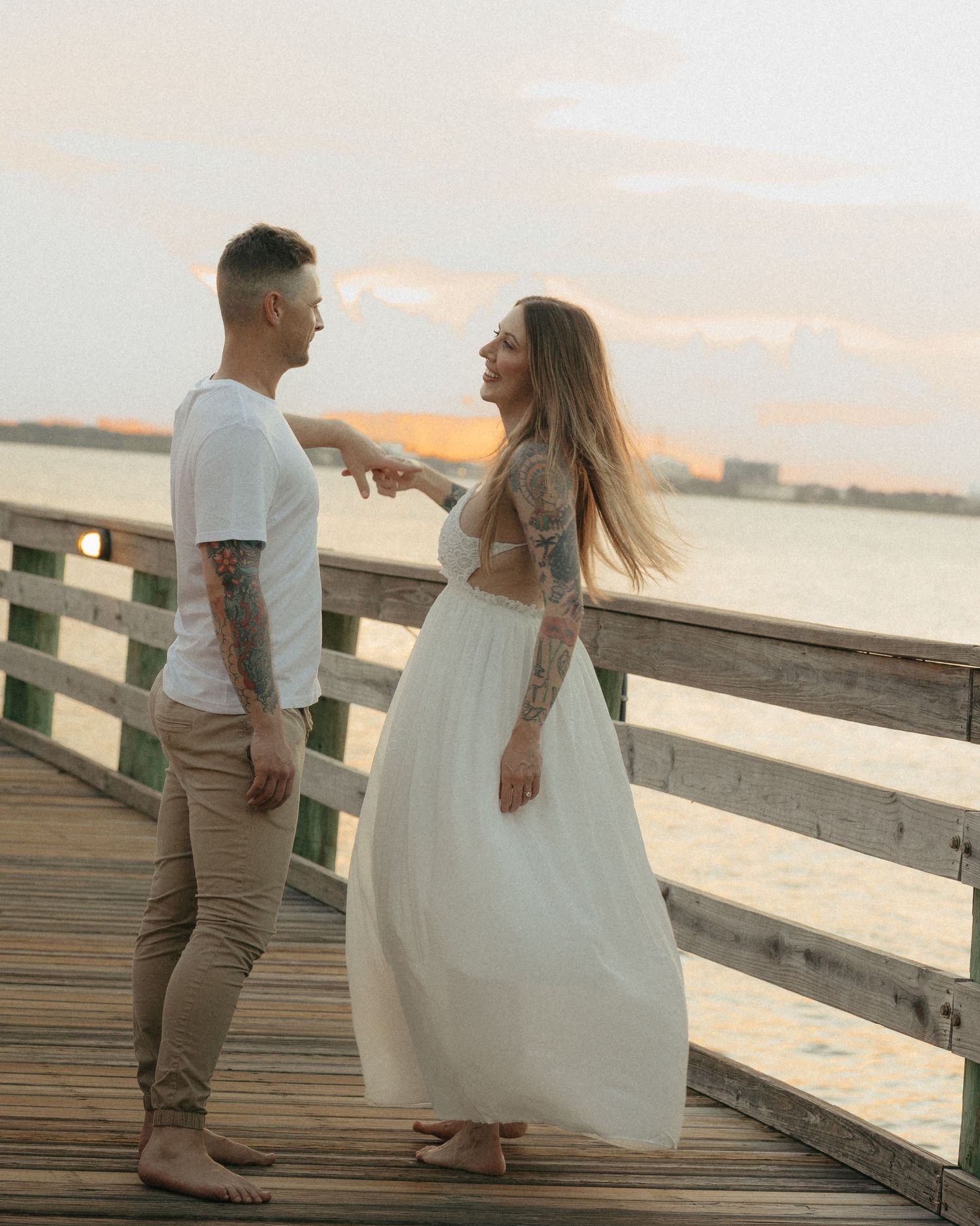 Couple in white outfits dancing on a wooden pier at sunset over the water.