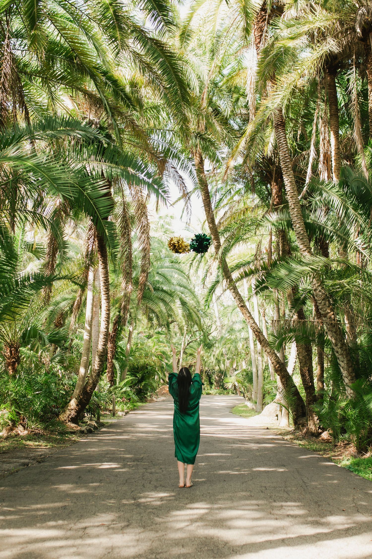 Woman in green dress standing on a path surrounded by tall tropical palm trees tossing a bouquet.