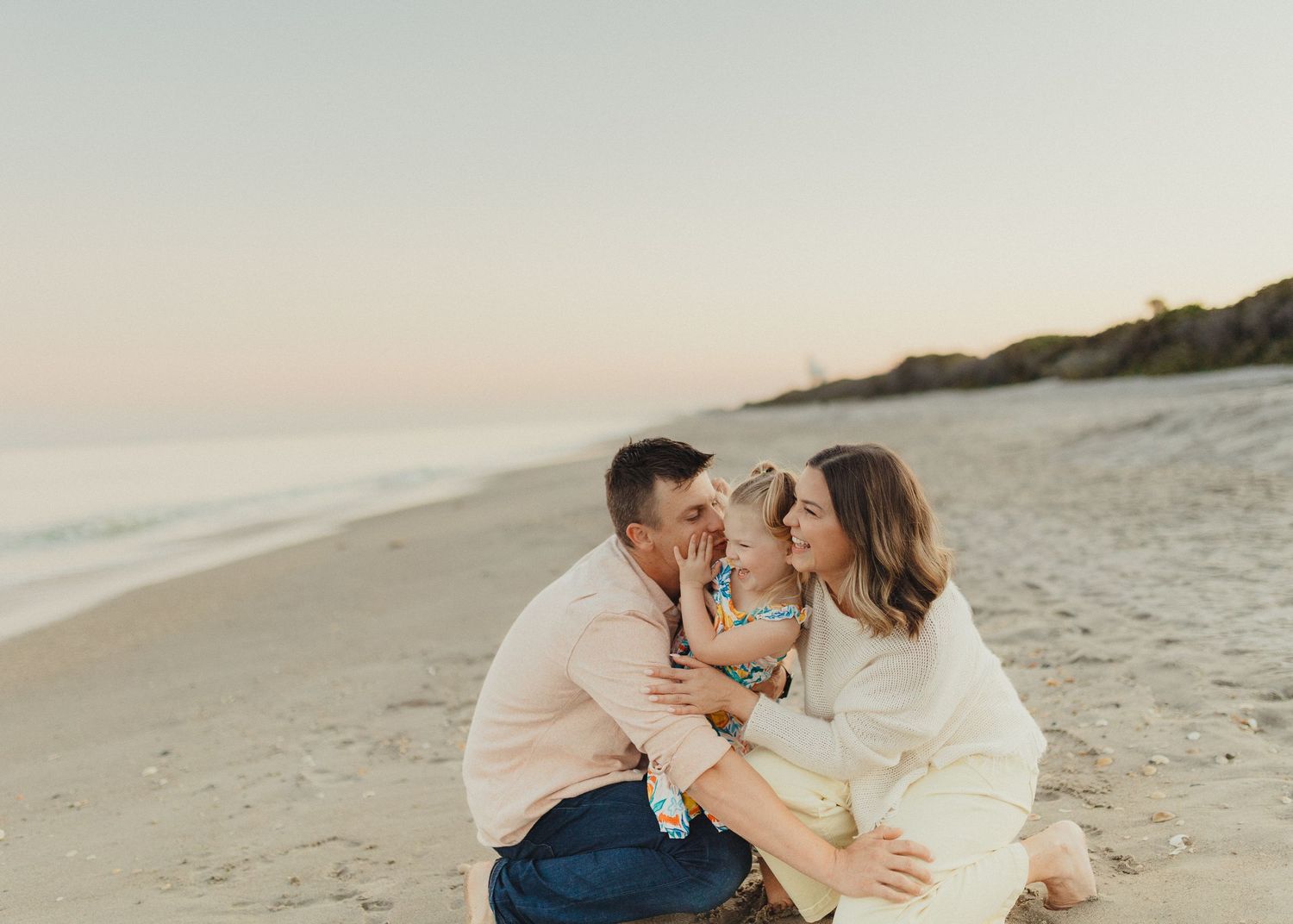 Family of three sharing a tender moment on a sandy beach at sunset, parents kissing child between them.