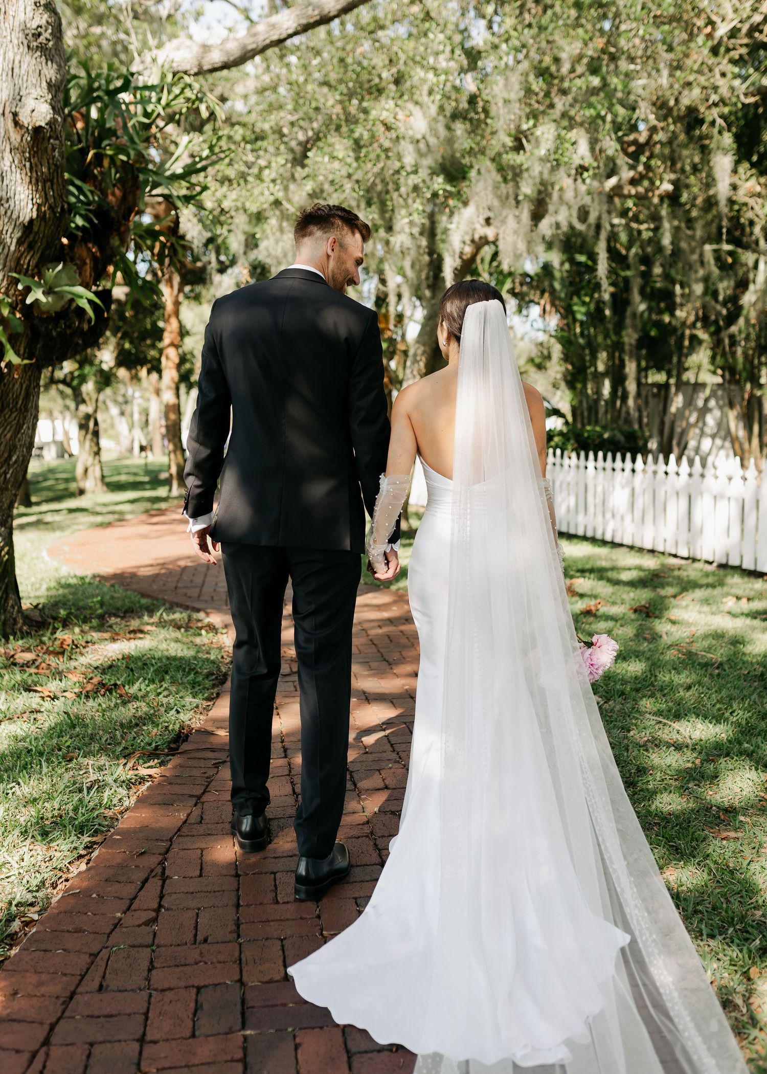 Bride and groom walking hand in hand down brick path through lush tree-lined garden.