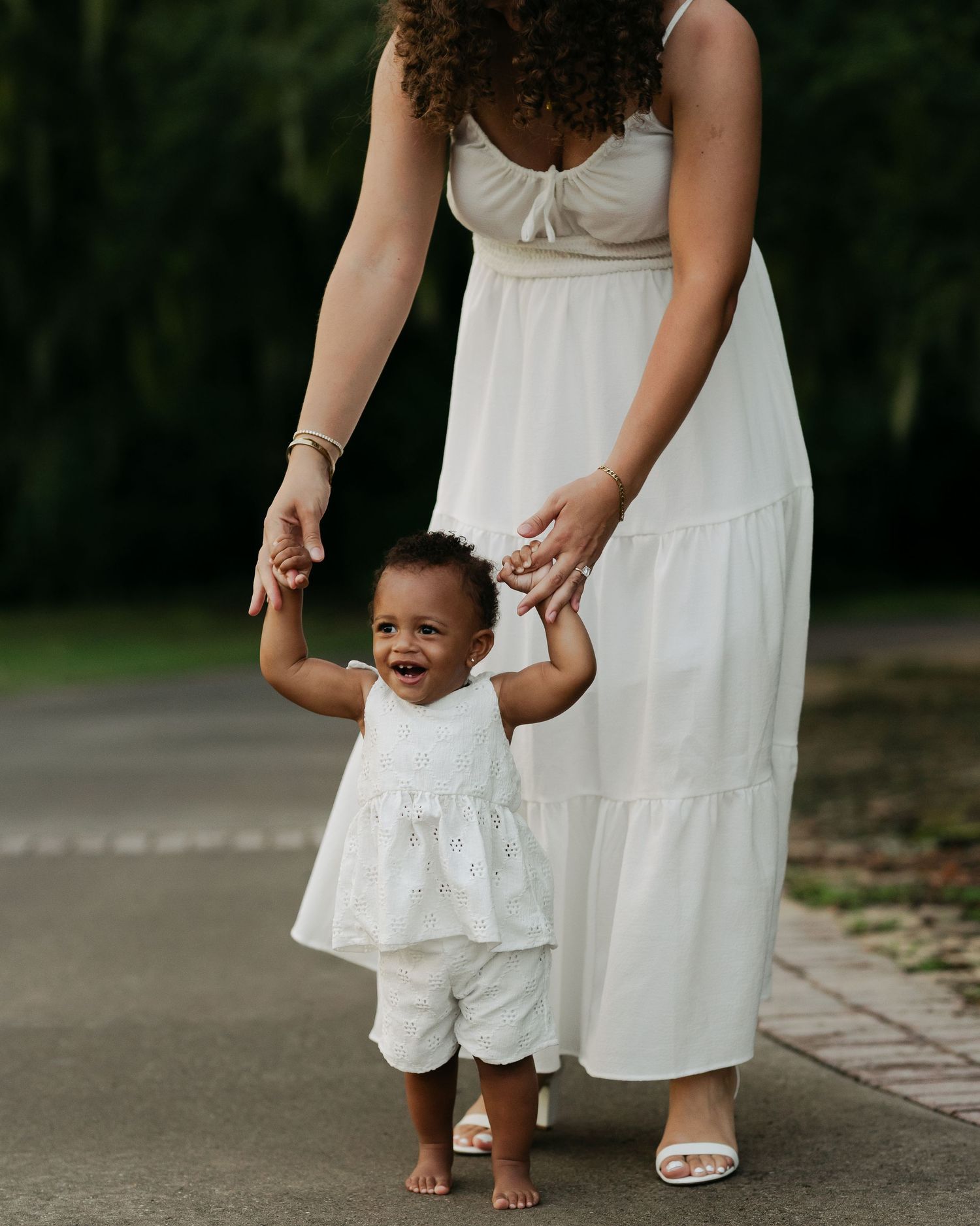 Adorable baby in white outfit learning to walk while holding mother's hands on an outdoor path.