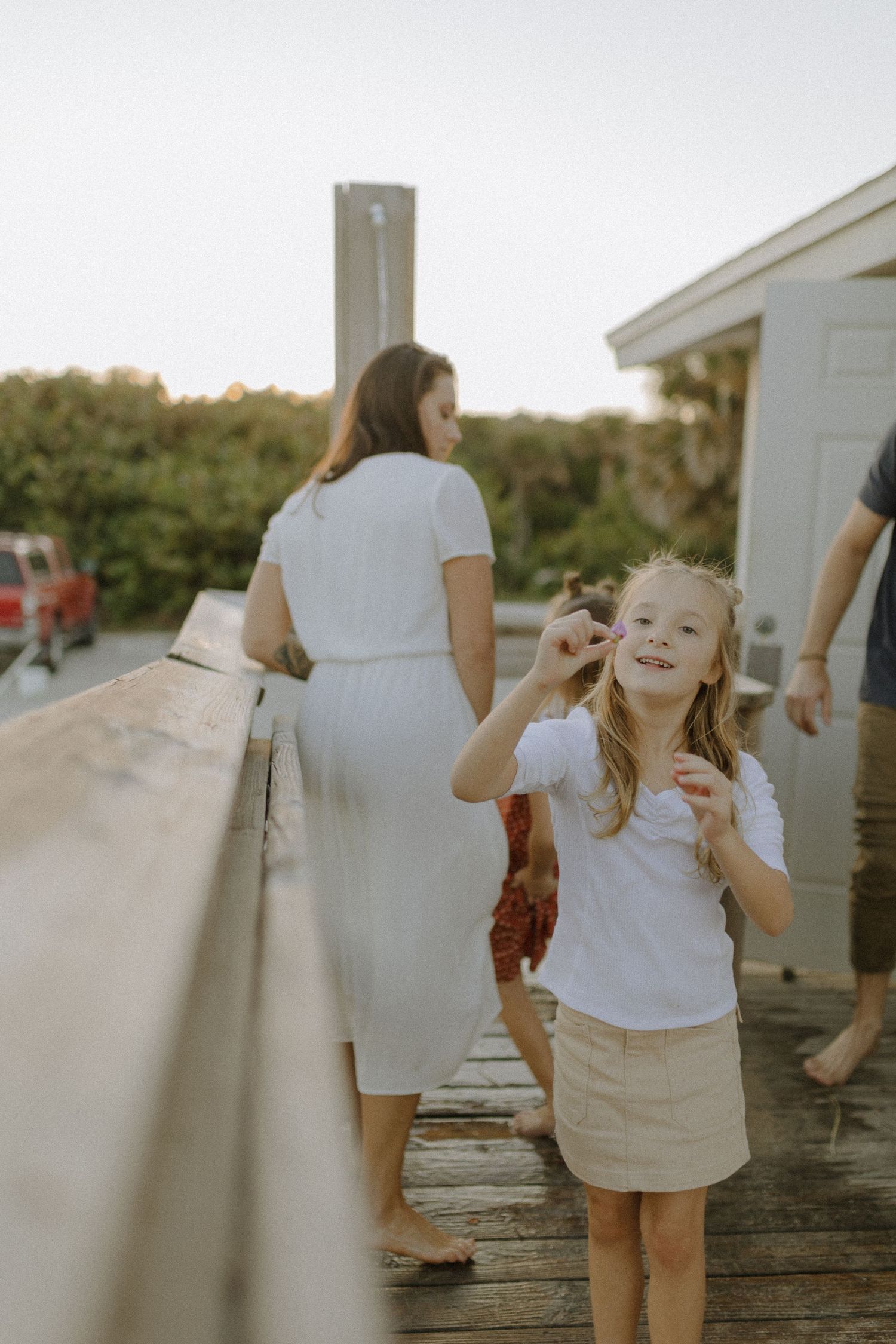 Young blonde girl smiling on wooden boardwalk at sunset with family members visible in background.