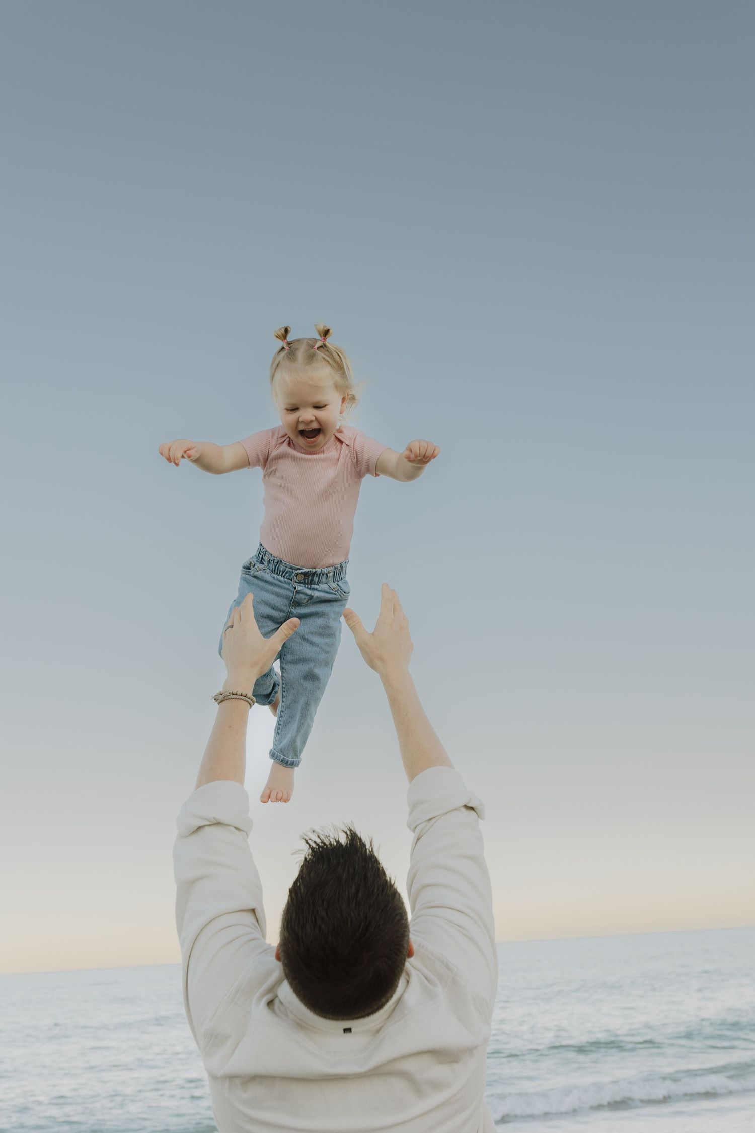Father tossing laughing baby girl into the air on a calm beach at dusk.