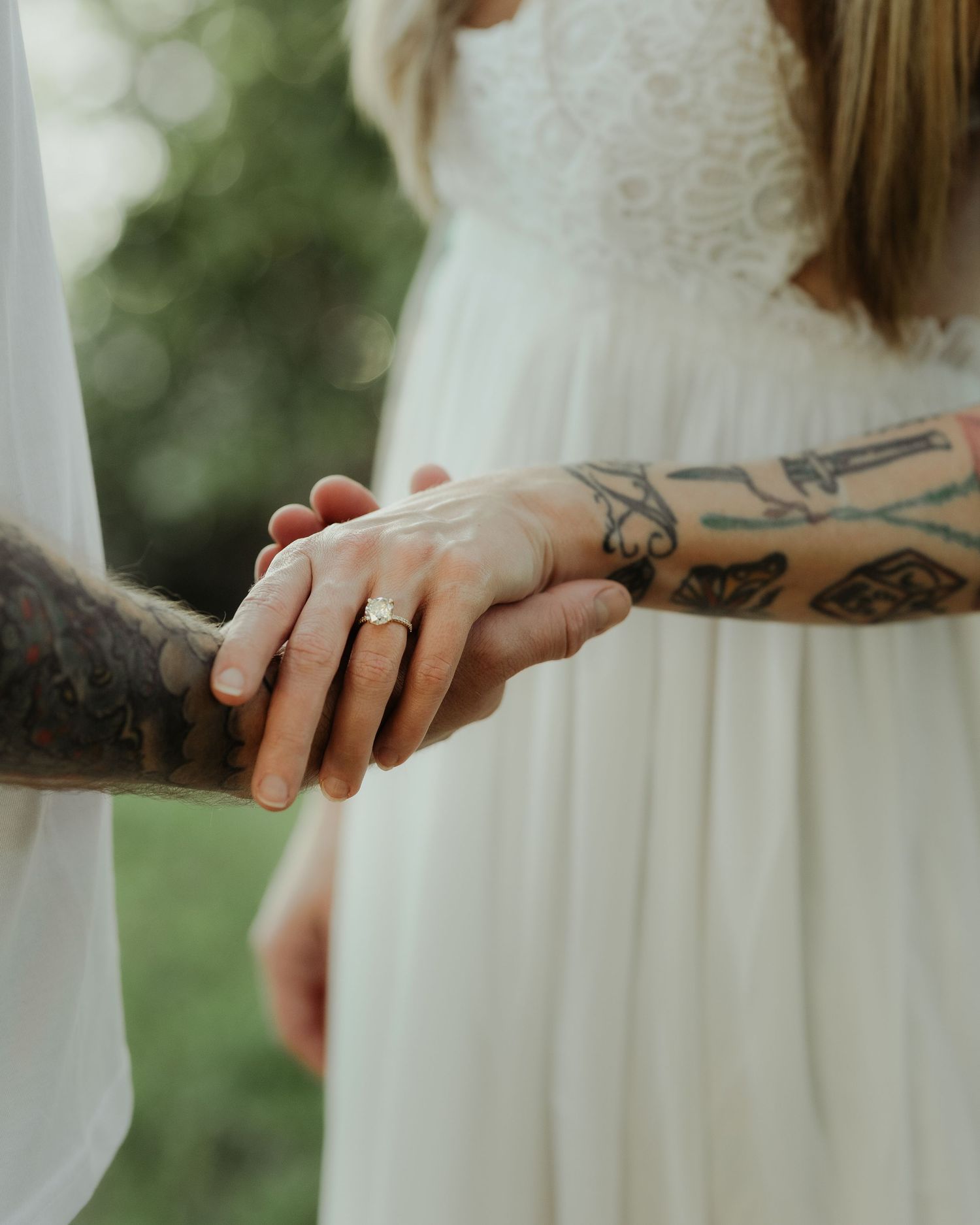Close-up of tattooed couple holding hands showing a gold engagement ring outdoors.