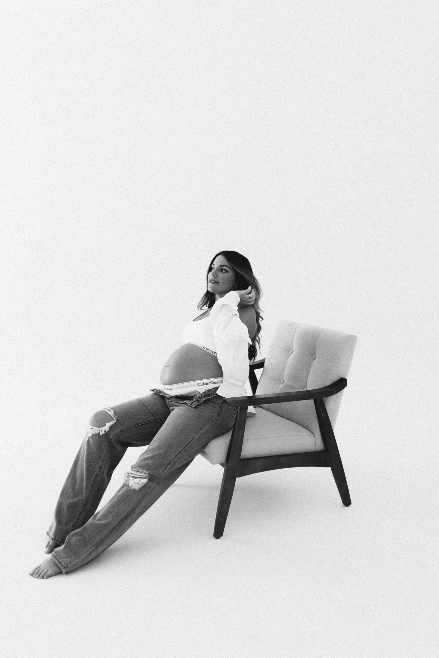 Black and white photo of a pregnant woman sitting on a chair in ripped jeans and white shirt.
