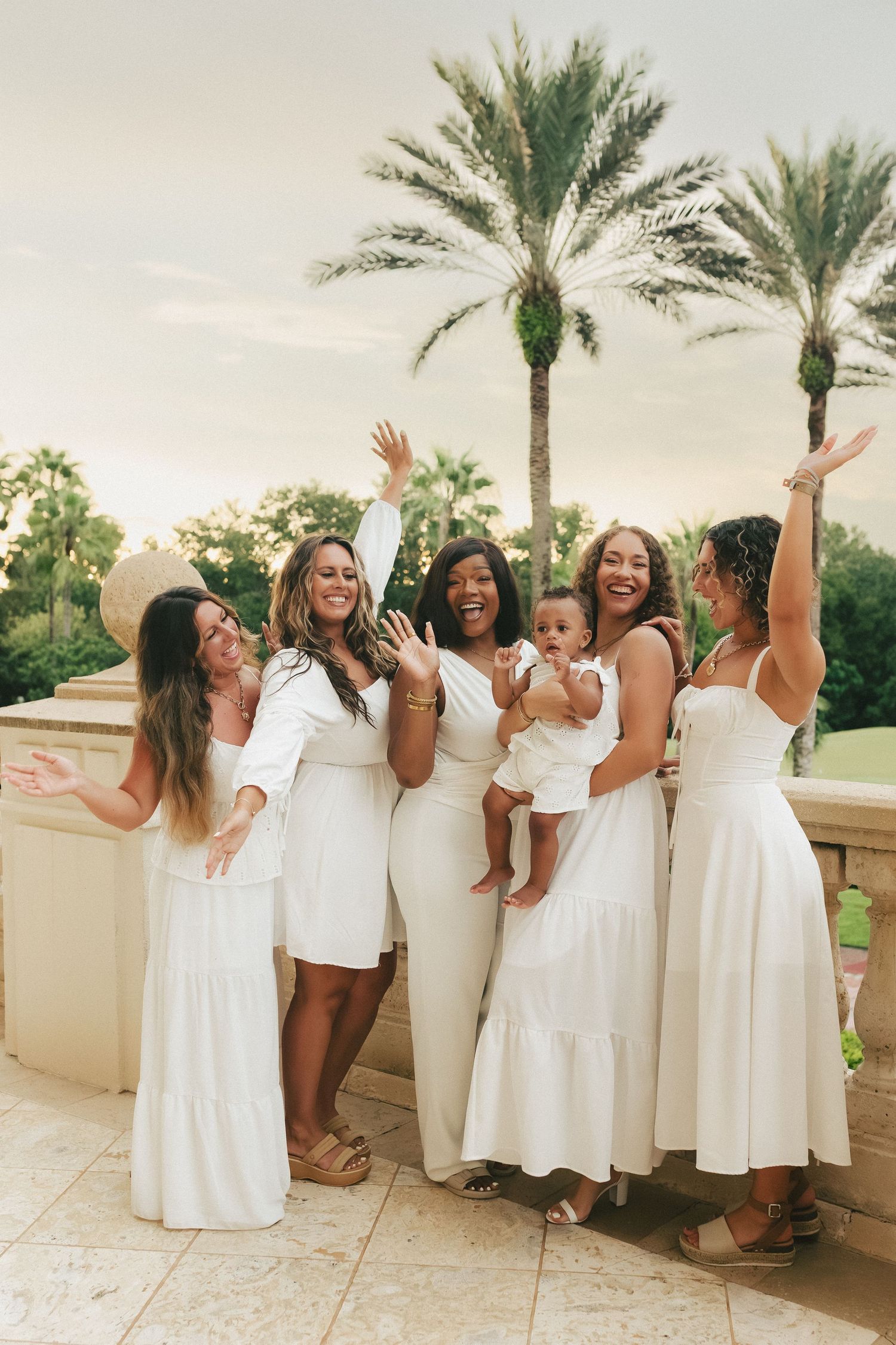 Five women in white dresses celebrating outdoors with palm trees, raising hands joyfully while one holds a baby.