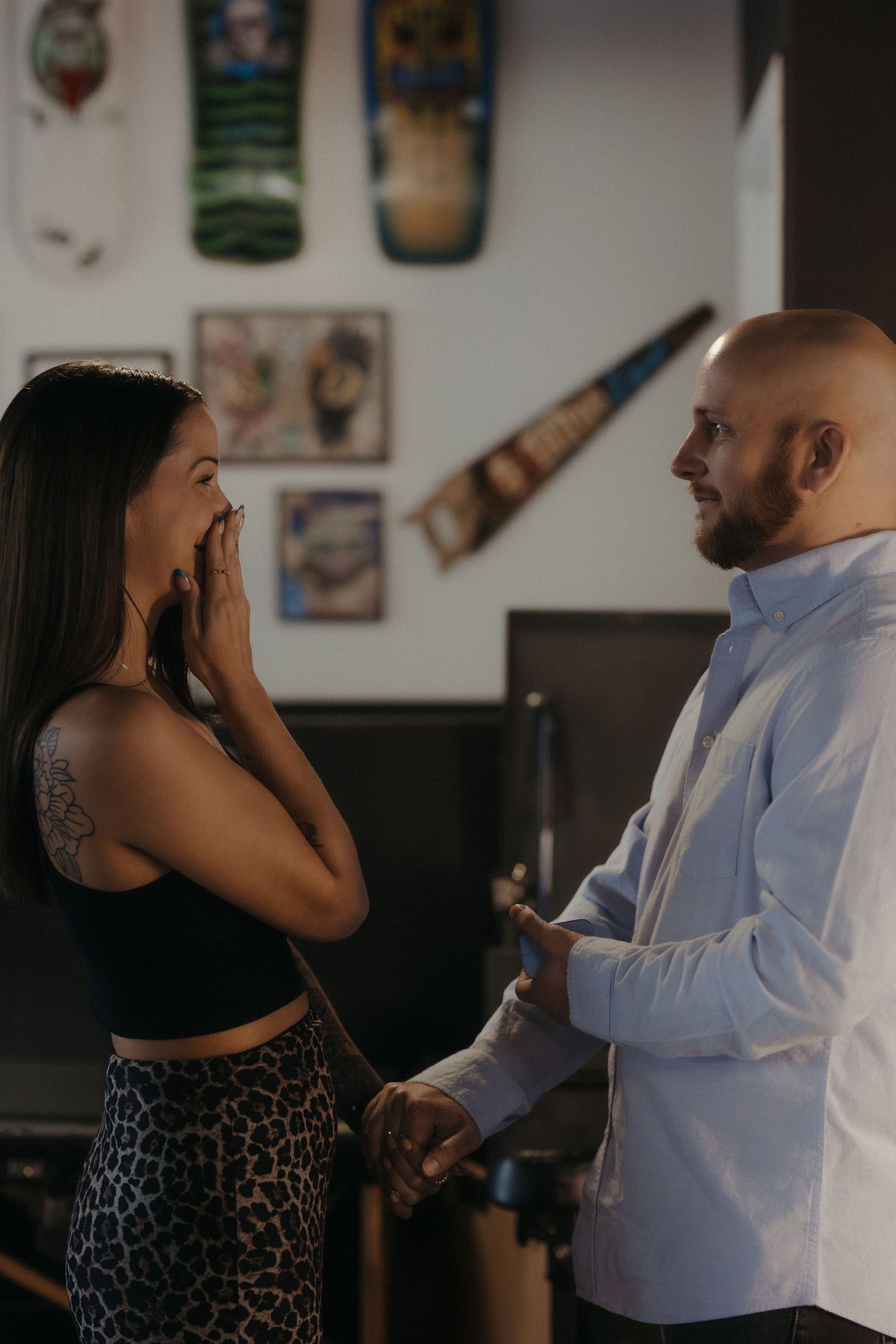 A man surprises a tattooed woman inside a decorated tattoo shop, she reacts with joy.