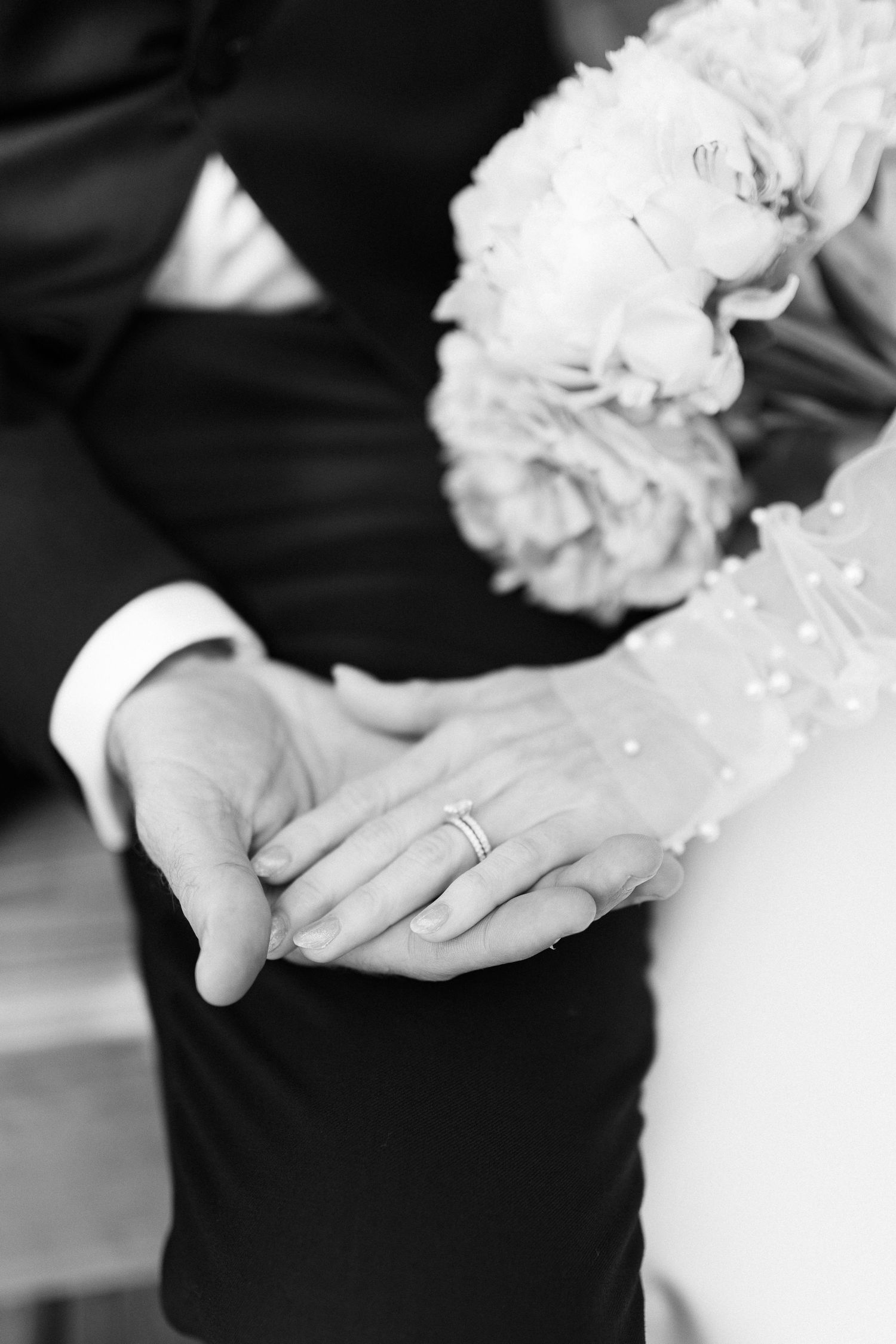 Close-up black and white photo of newlyweds holding hands showing wedding rings and bridal bouquet.