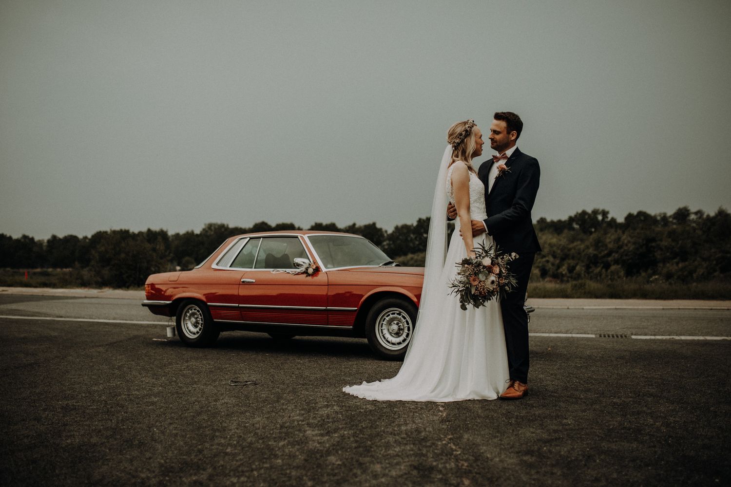 Brautpaar steht vor einem roten Oldtimer, lacht sich an, umgeben von Abendlicht – emotionale Hochzeitsfotografie in Zeven von Ann-Kathrin Br