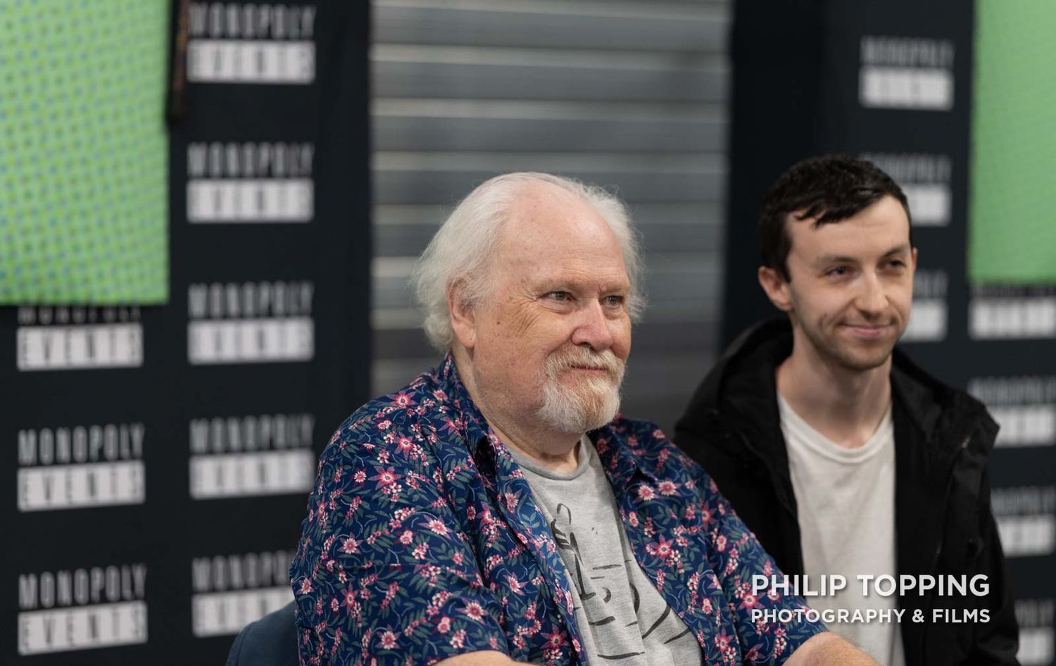 Colin Baker is sitting down with an assistant beside him, chatting to someone.