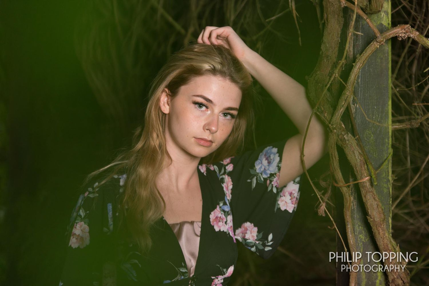 A female with her hand on the side of her head against a tree with lighting from a softbox off camera.