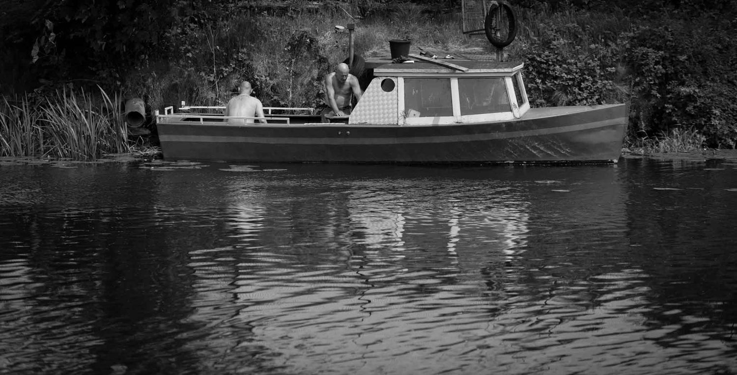 Two people are working casually on a small boat at the side of the river.