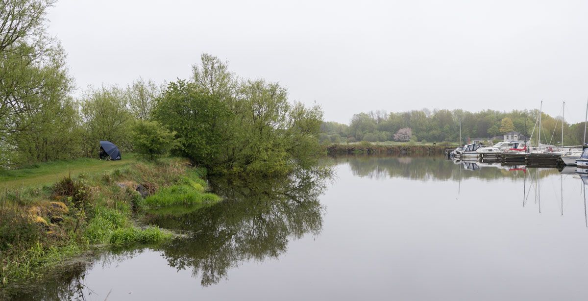 A person fishing under an umbrella with boats in the distance right.