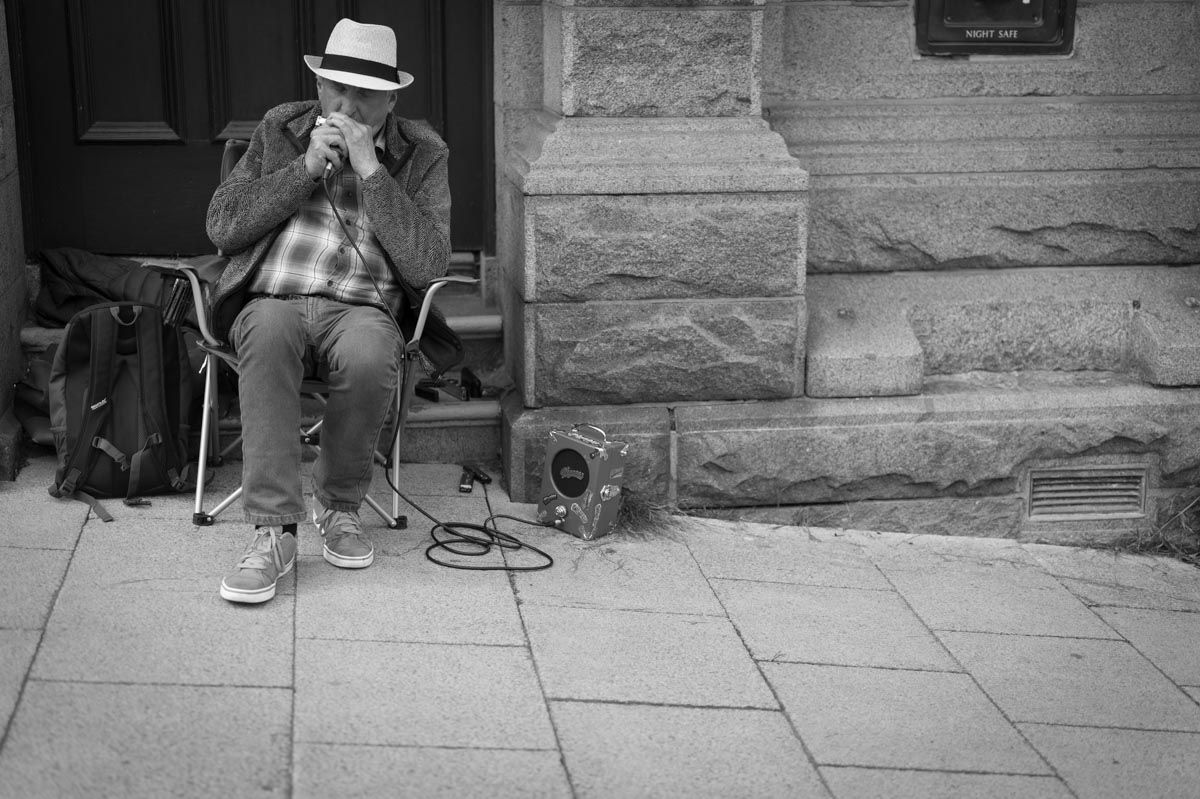 An older man is sitting down playing a harmonica with a very small speaker beside him.