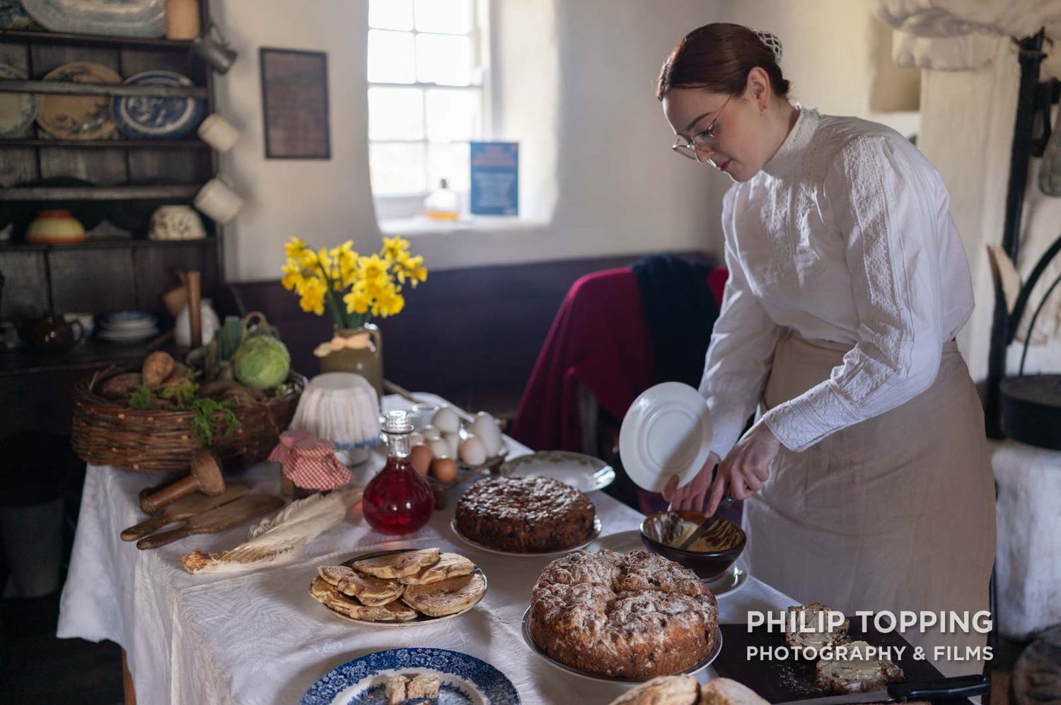 Wide photo of a female dressed in Victorian clothes cutting bread on a table.