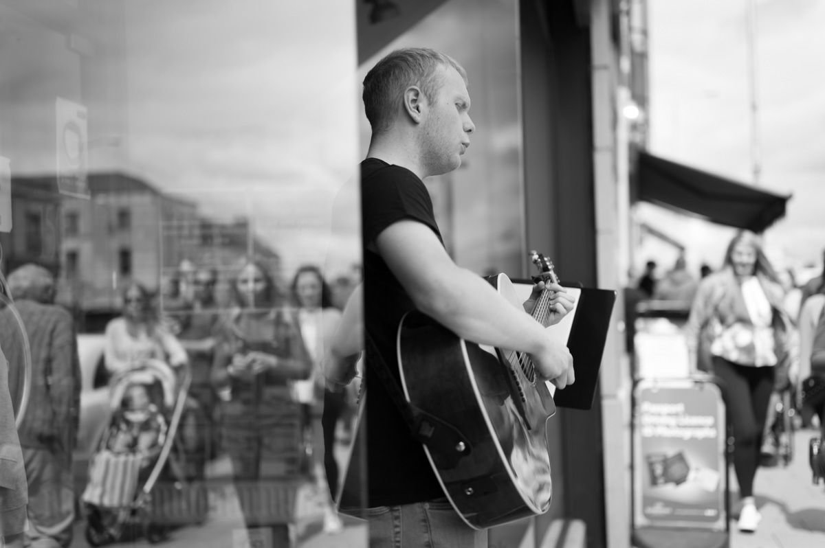 Guitarists playing at the entrance of the shop with reflections of people walking by.