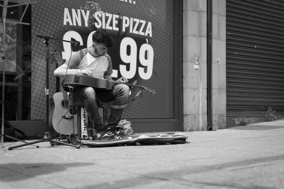 A busker playing a slide guitar is sitting down.