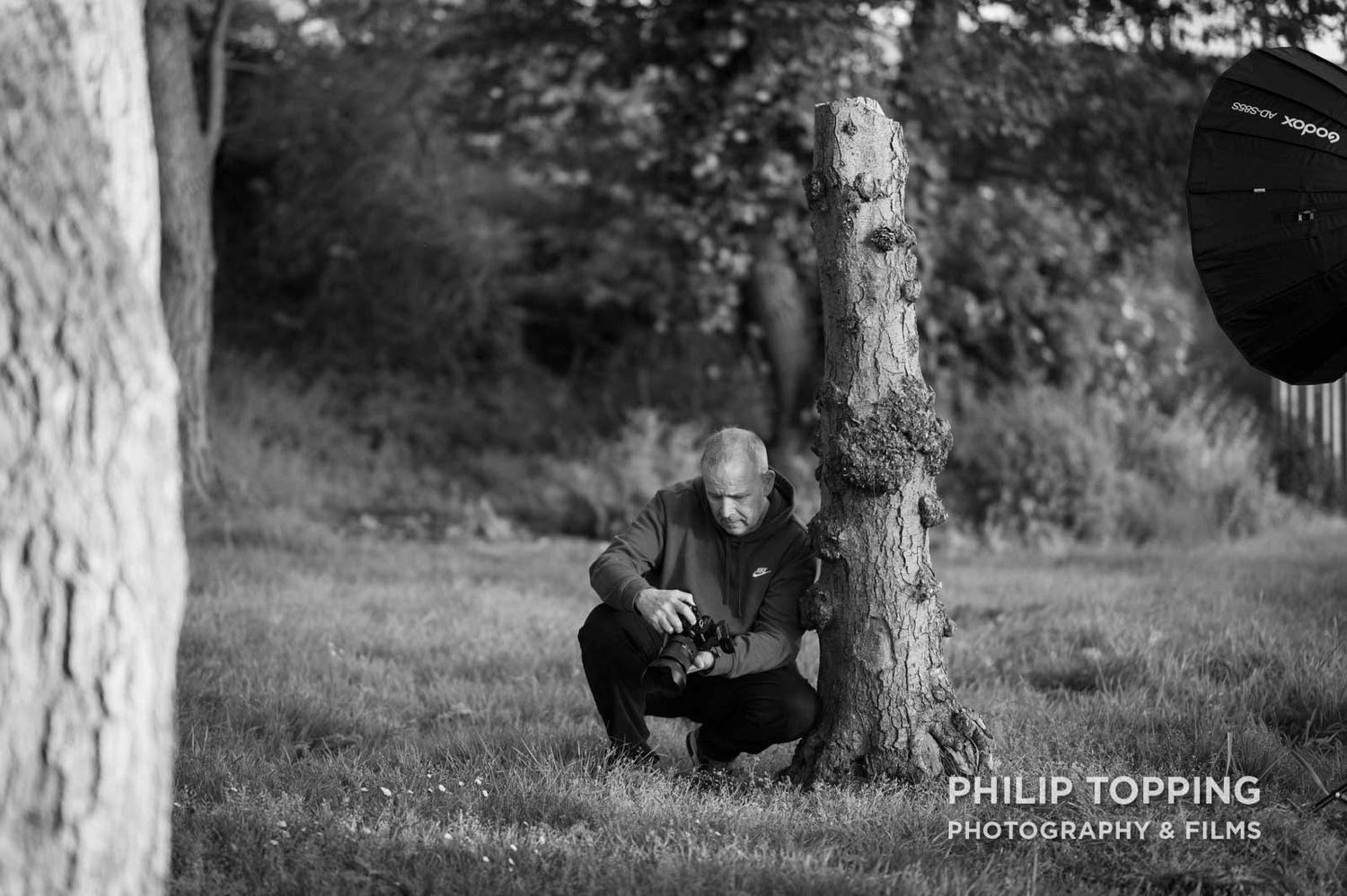 Club member crouching beside a tree in Hoys Meadow, checking images on his camera in mono.