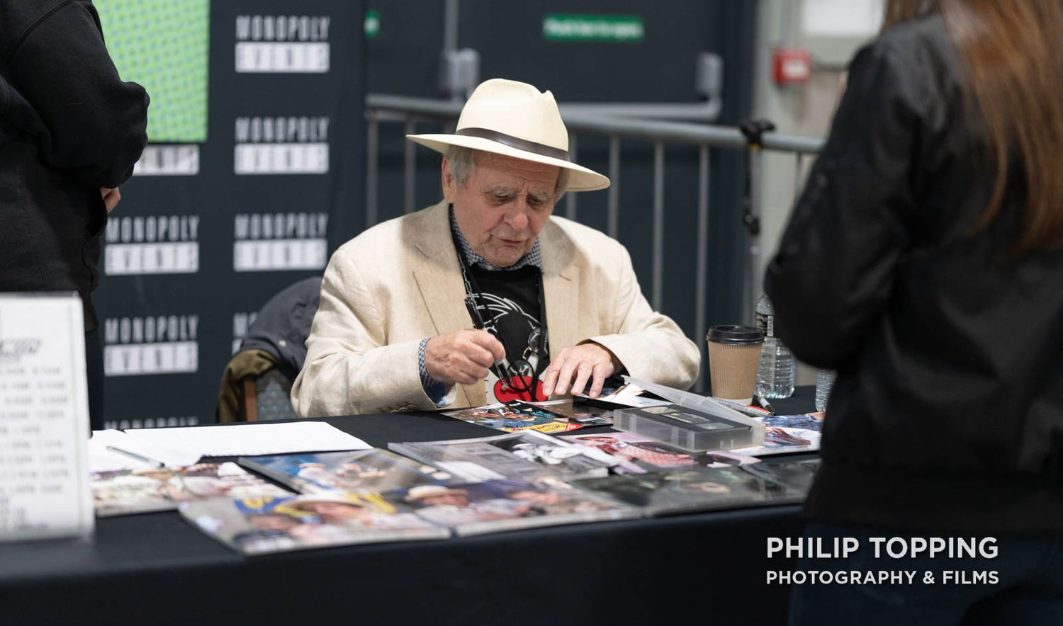 Celebrity guest Sylvester McCoy is sitting down at his table, signing autographs.