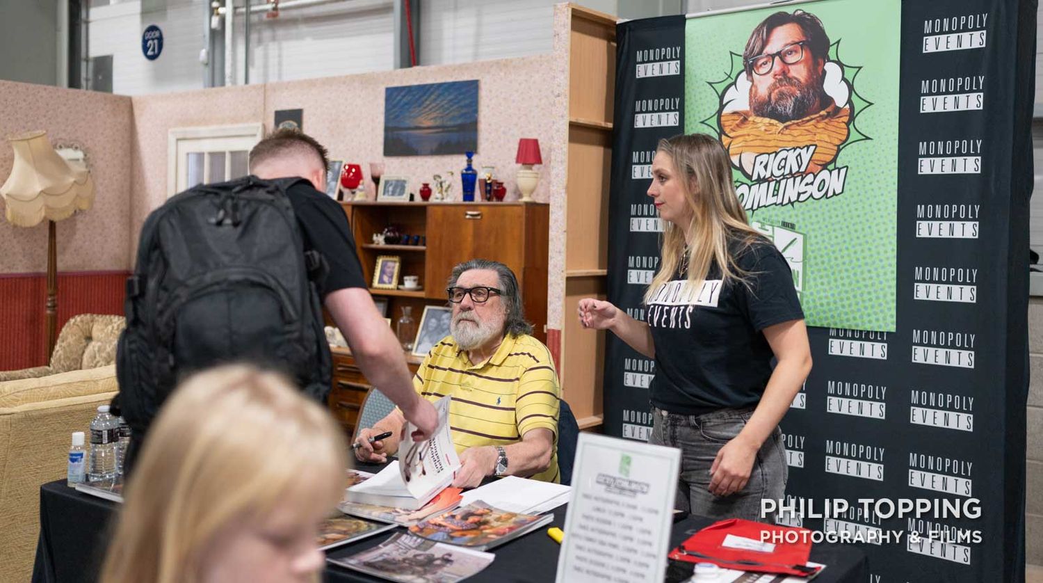 Ricky Tomlinson is sitting at his table with a yellow shirt on, talking to someone.