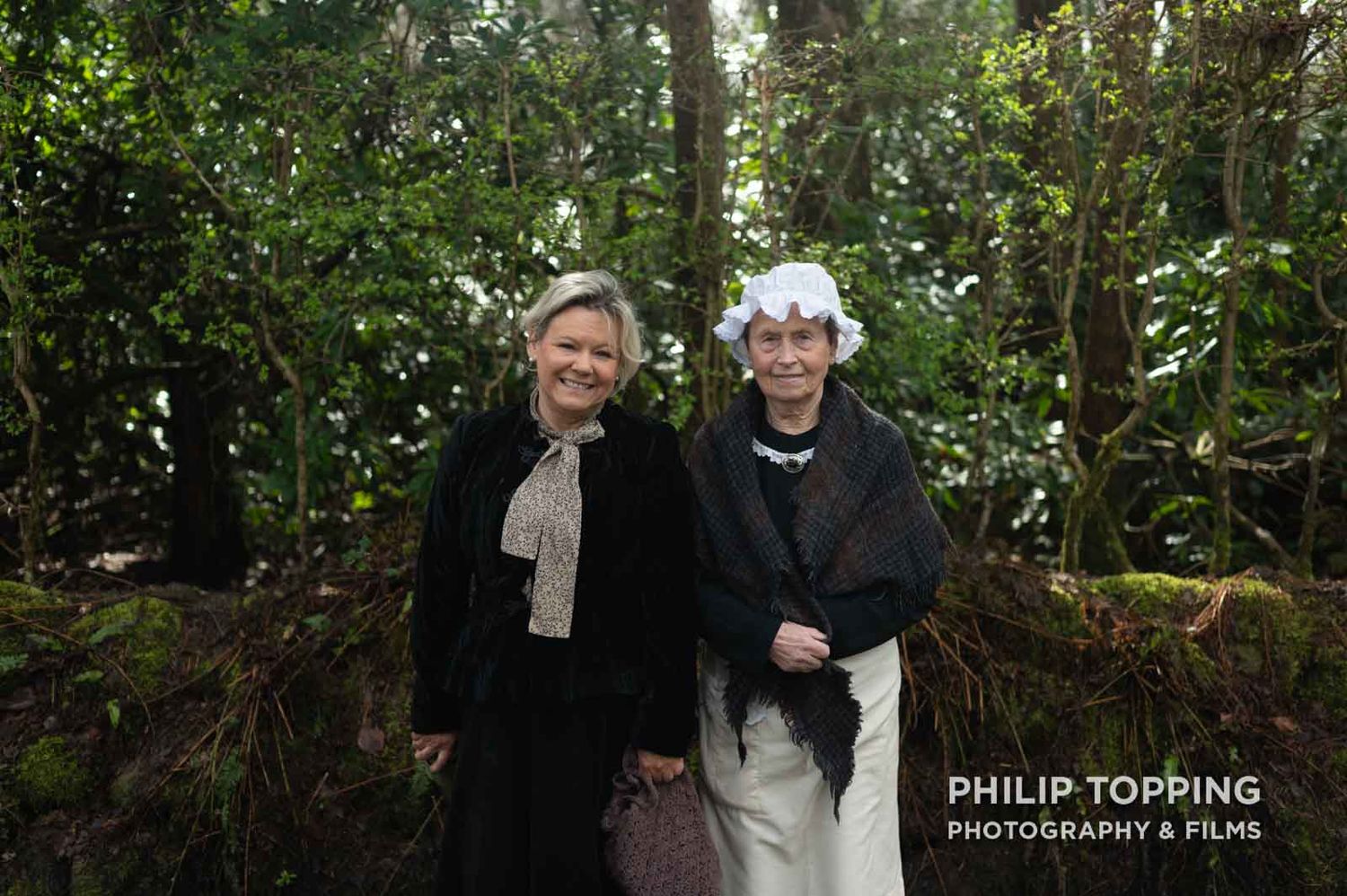 Two females dressed up as historical interpreters are looking at the camera in front of trees.