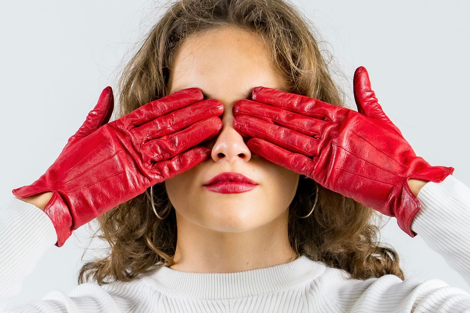 Woman with curly hair covering eyes with red leather gloves wearing white sweater.