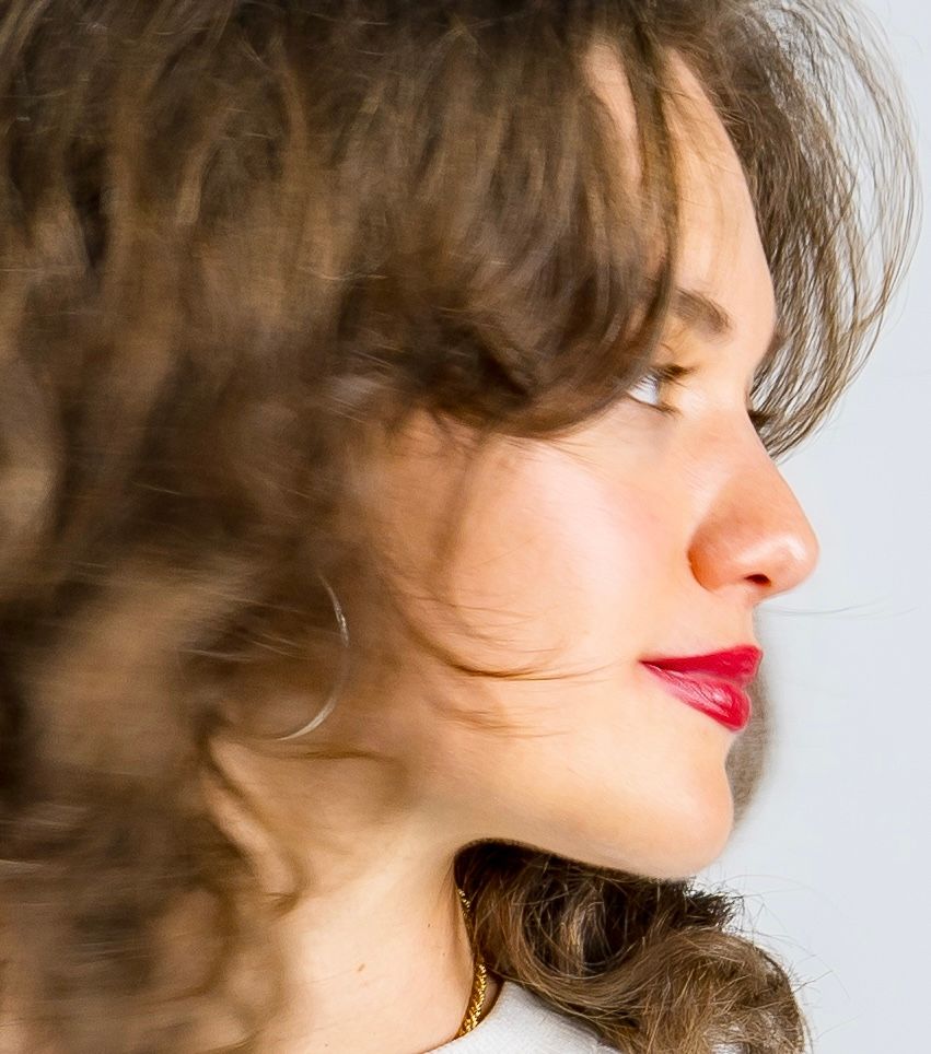 Side profile of woman with curly brown hair and red lips against bright background.