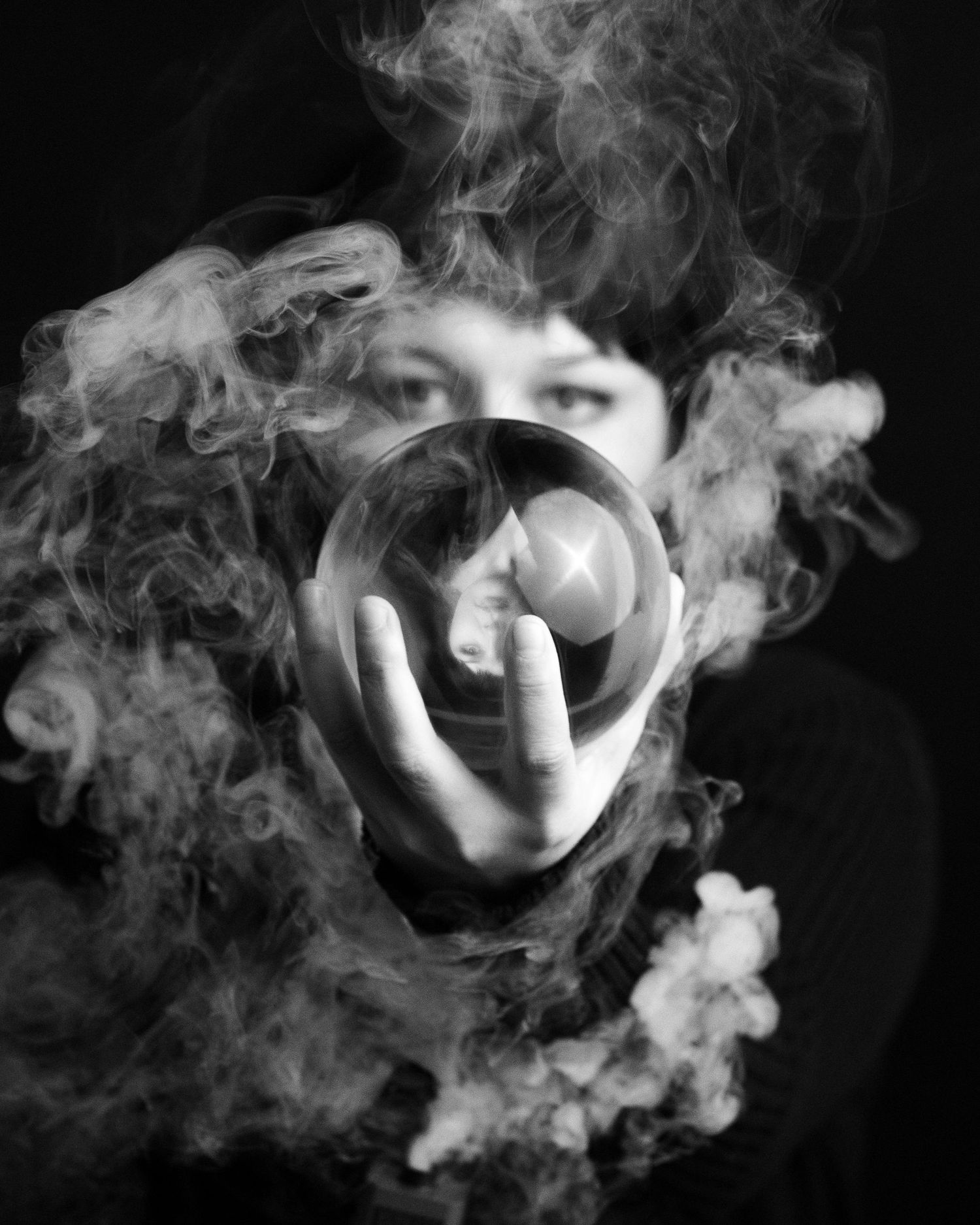 Black and white photo of hands holding crystal ball with smoke swirling around in dark setting.