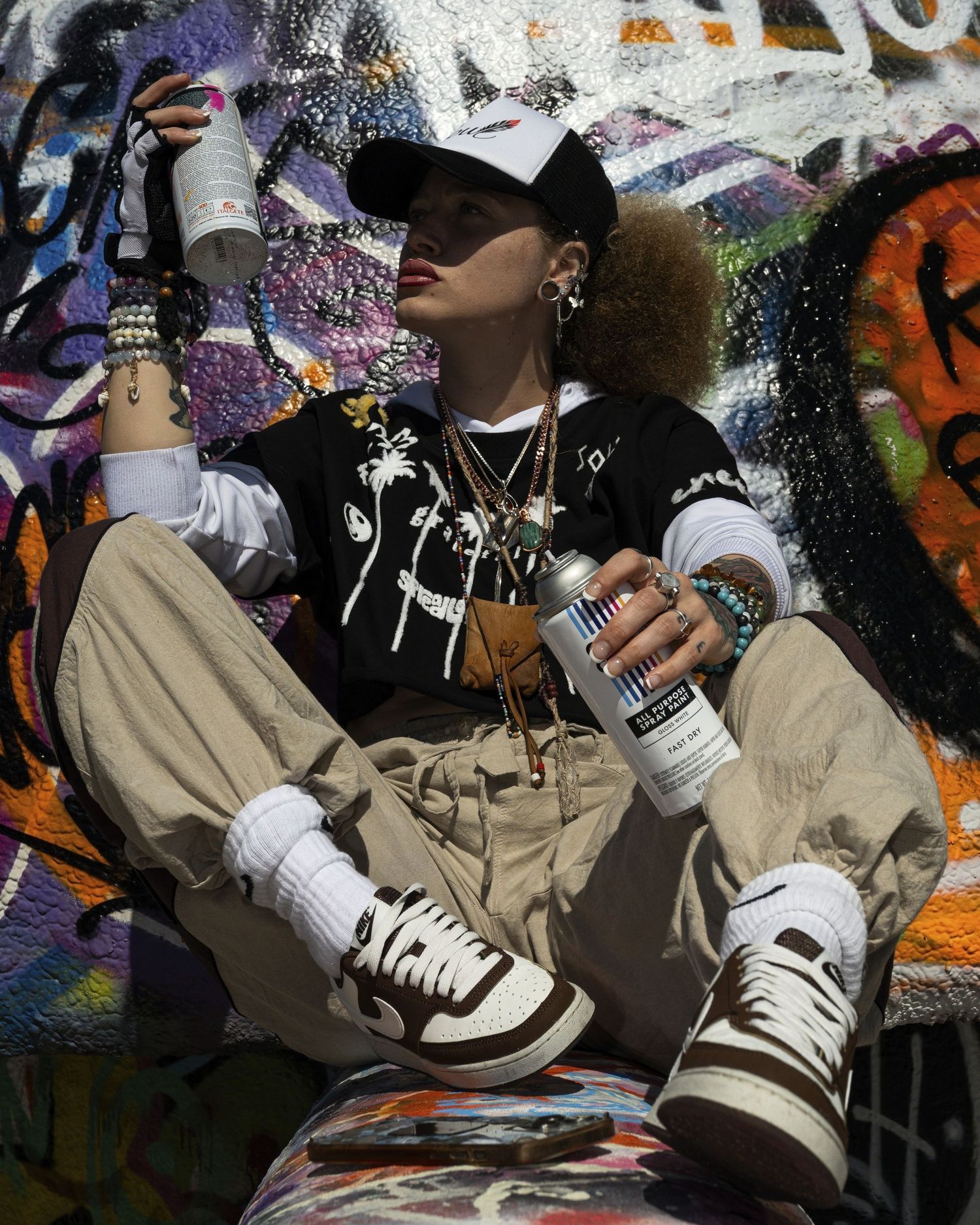 Young woman in streetwear poses with spray paint cans against a colorful graffiti wall.
