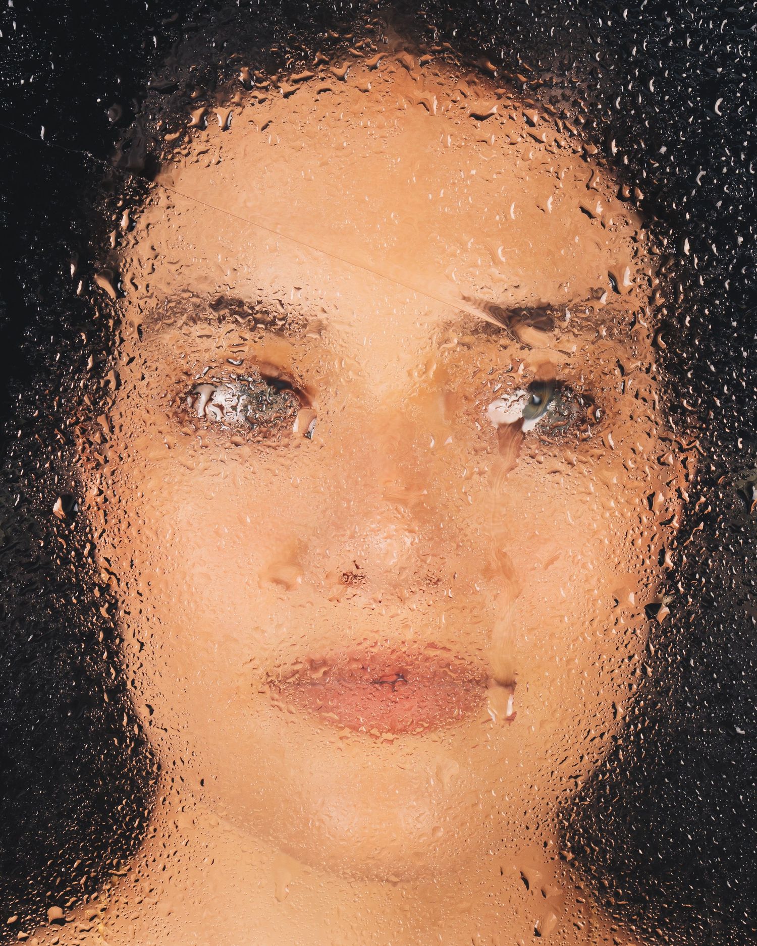 Woman's face viewed through wet glass covered in water droplets against dark background.