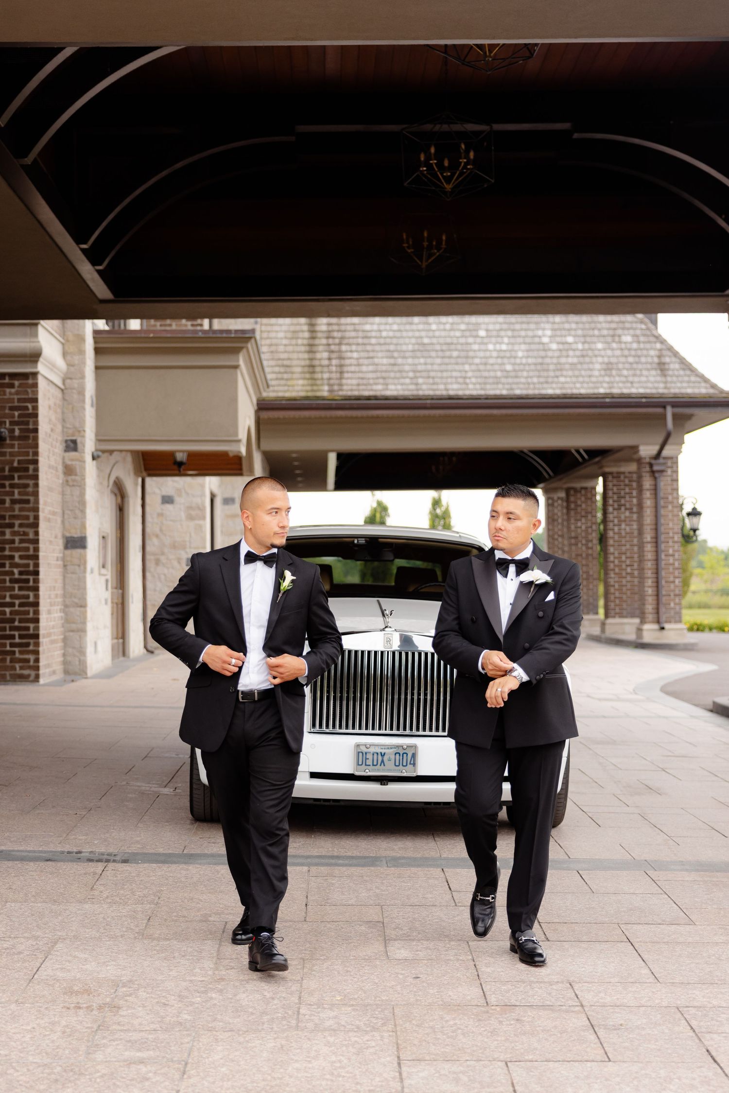 Two people in matching black tuxedos perform synchronized martial arts kicks in a covered outdoor area.