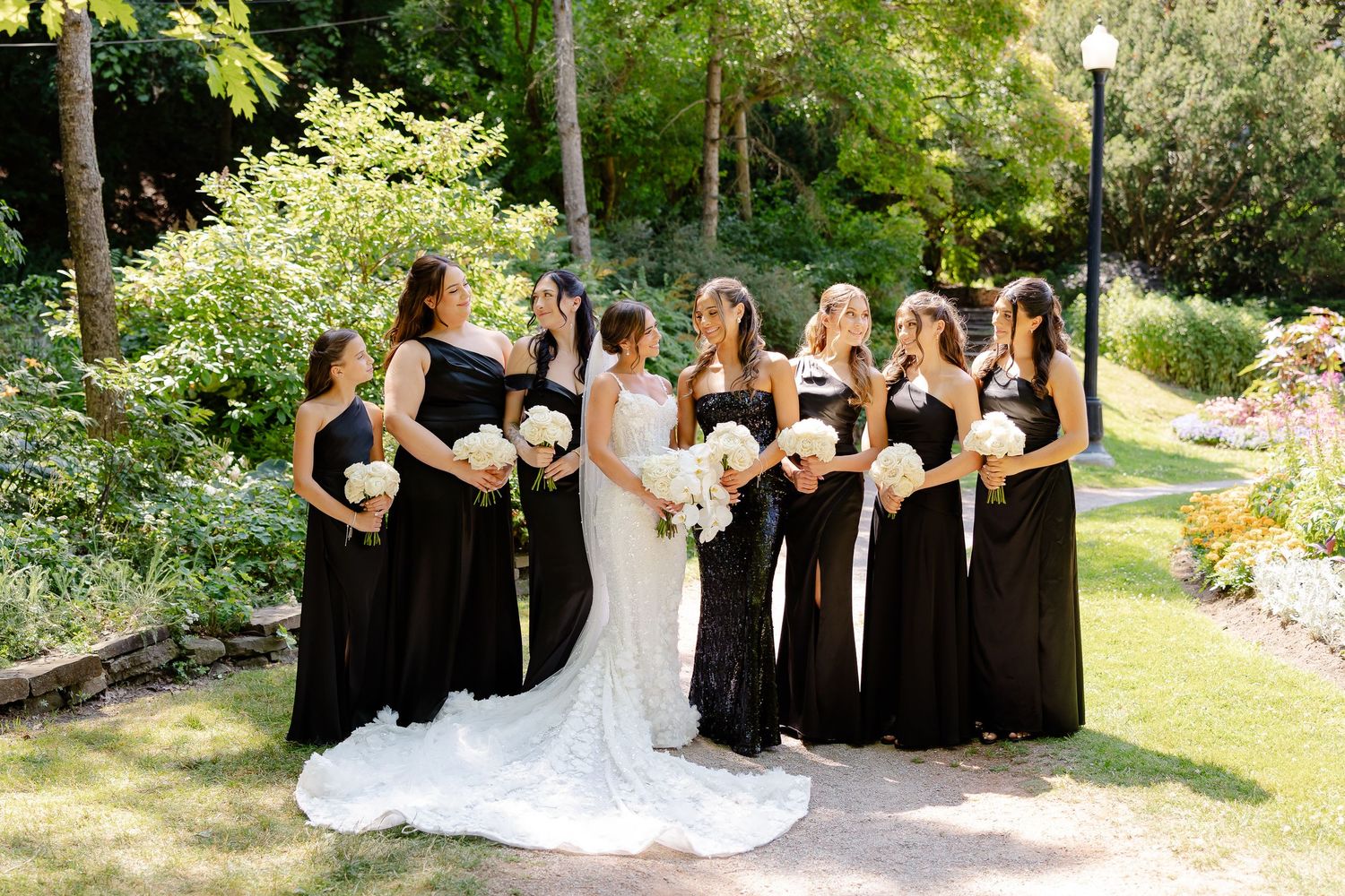 A bridal party in black gowns and a white wedding dress pose together in a sunlit garden setting.