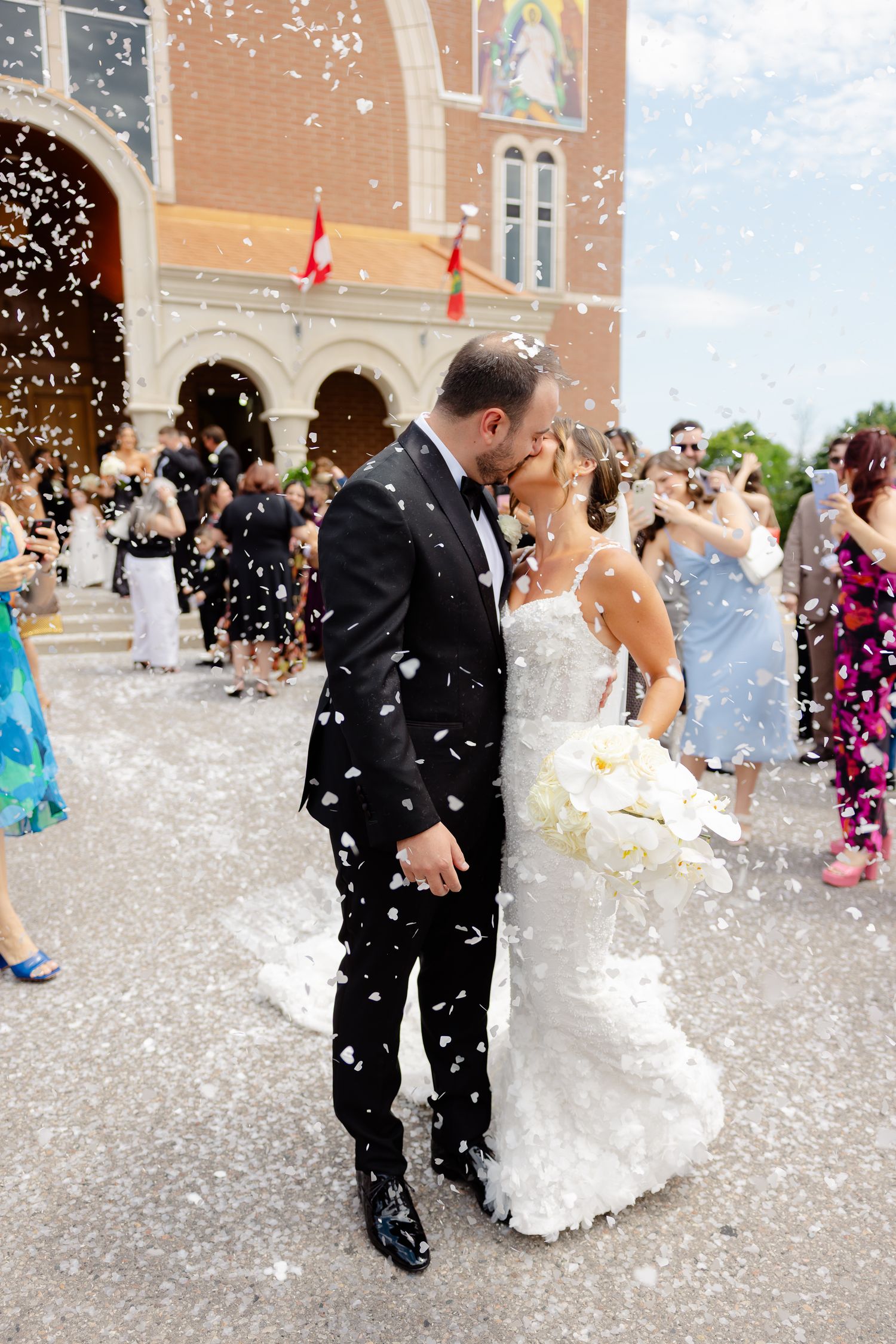 Newlyweds share a romantic kiss on church steps while guests shower them with white confetti on their wedding day.