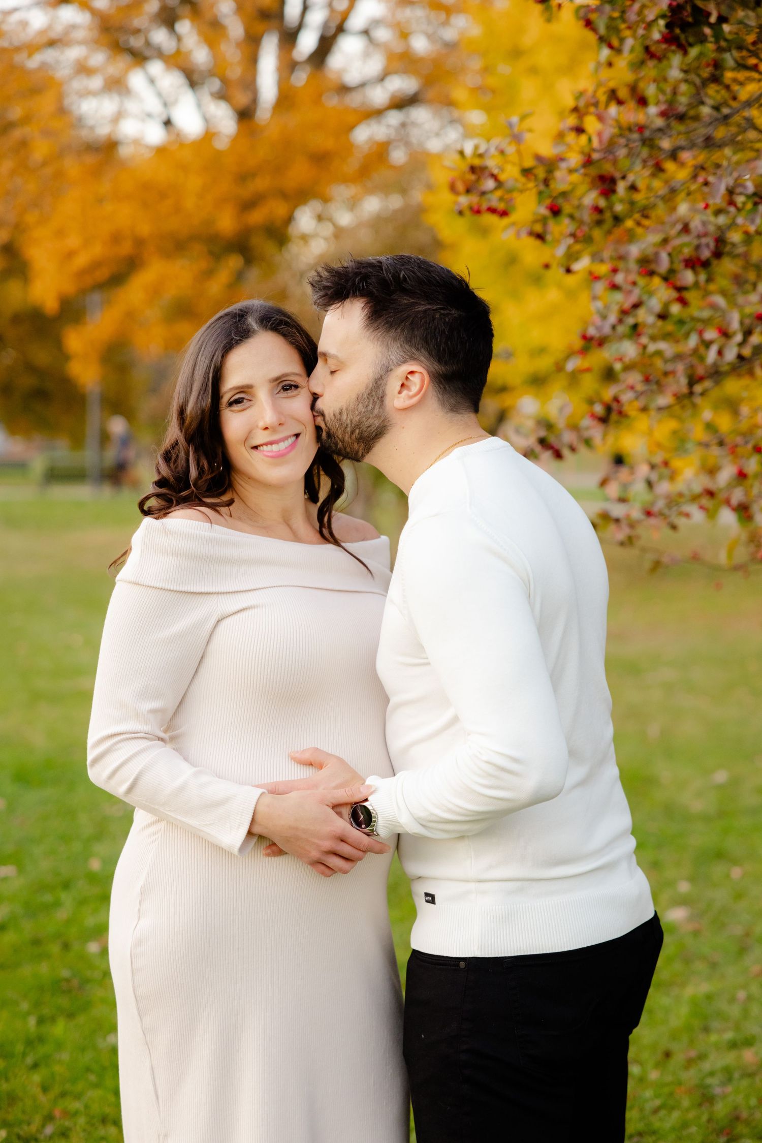 Couple in white sweaters hugging and smiling in fall park with colorful trees.