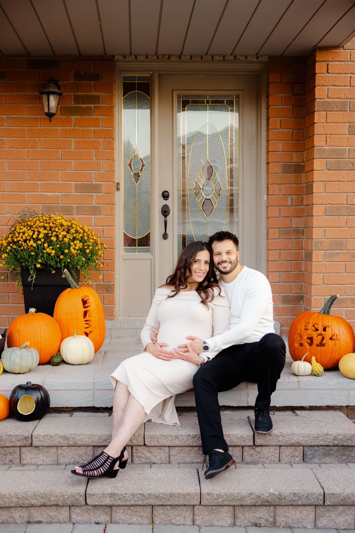A couple sits on steps surrounded by pumpkins and autumn decor in front of a brick house.