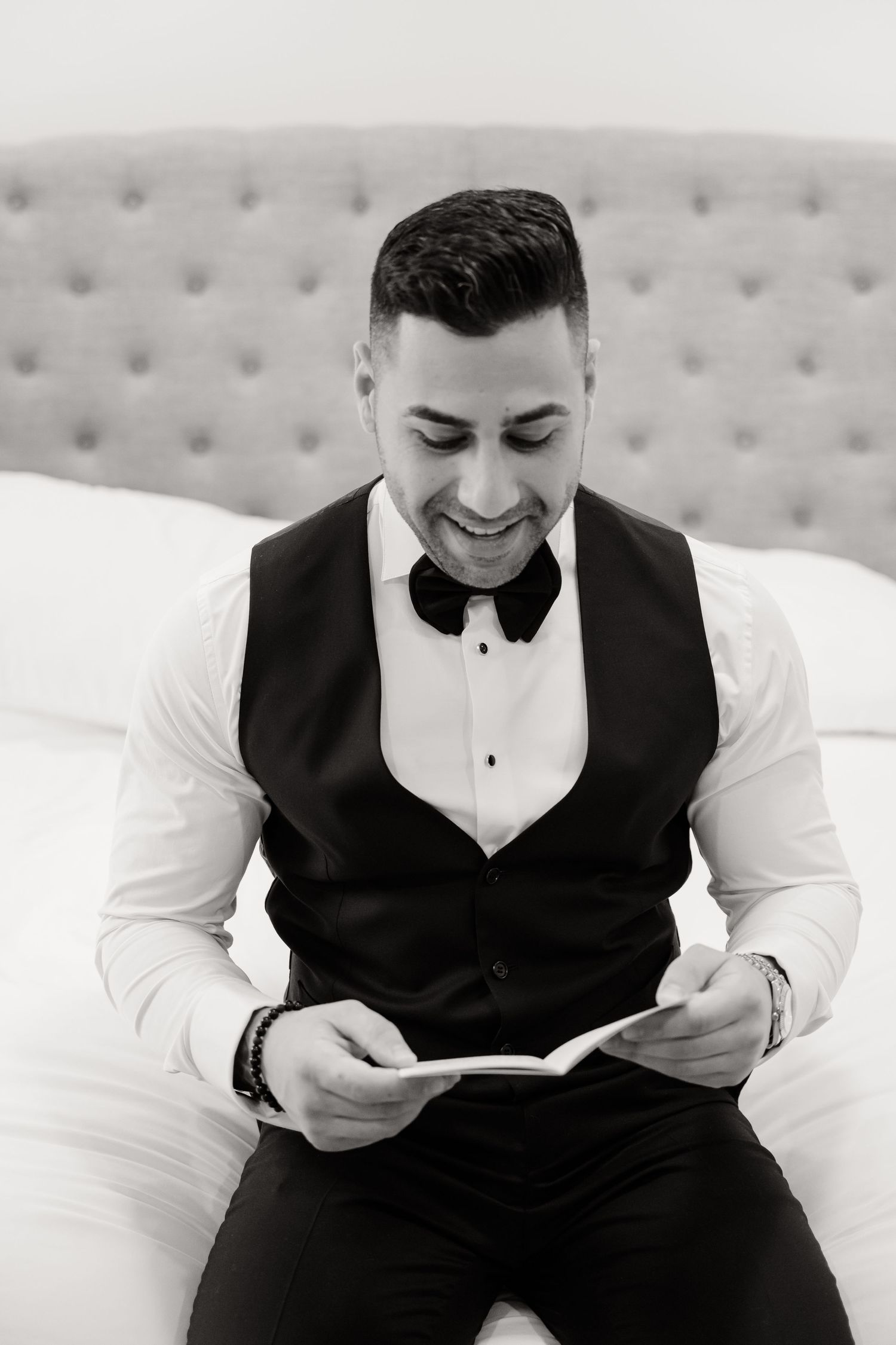 Person in black vest and bowtie smiling while reading a note on a tufted headboard bed.