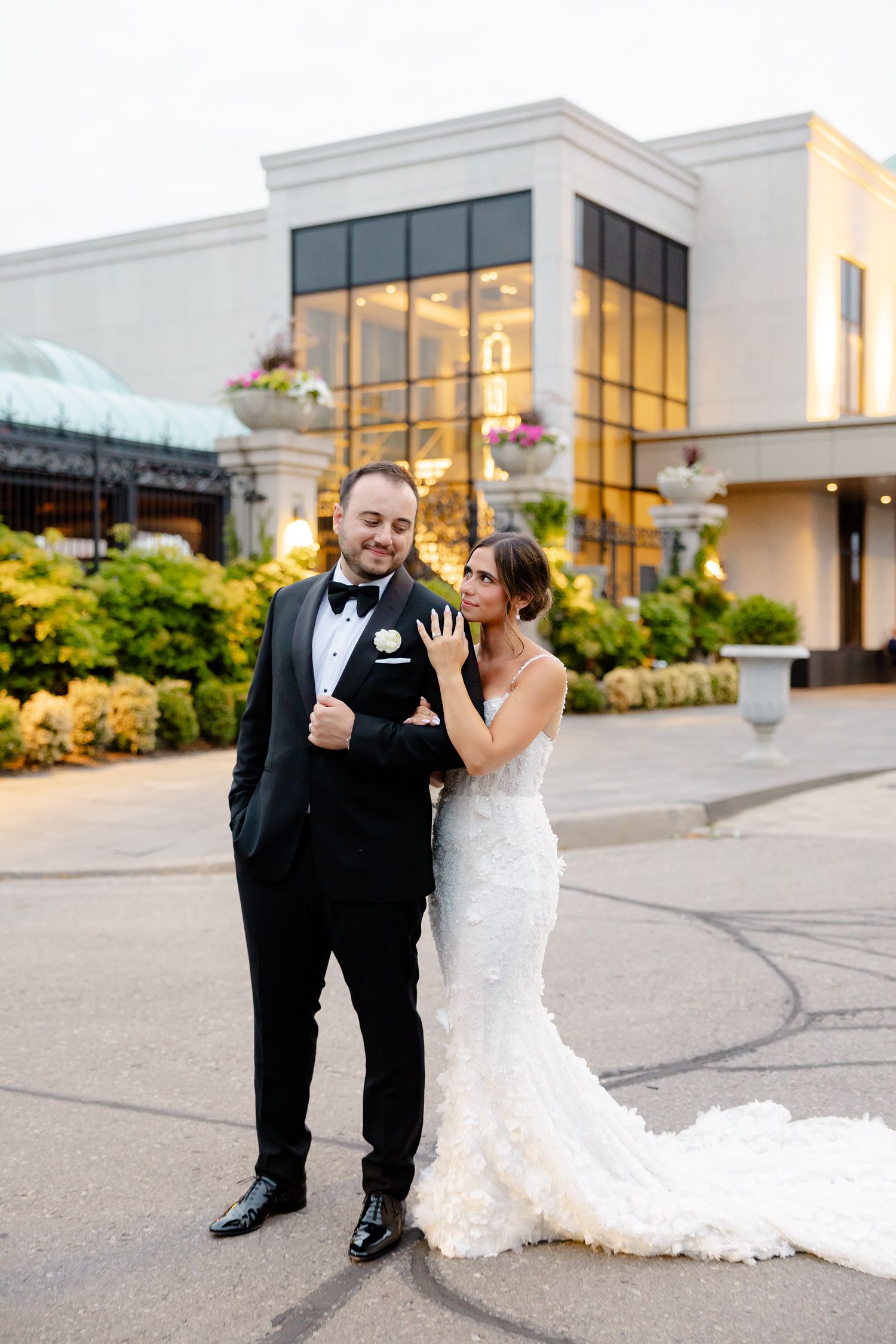 A couple shares a joyful moment outside an elegant venue with white architecture and floral decorations at sunset.