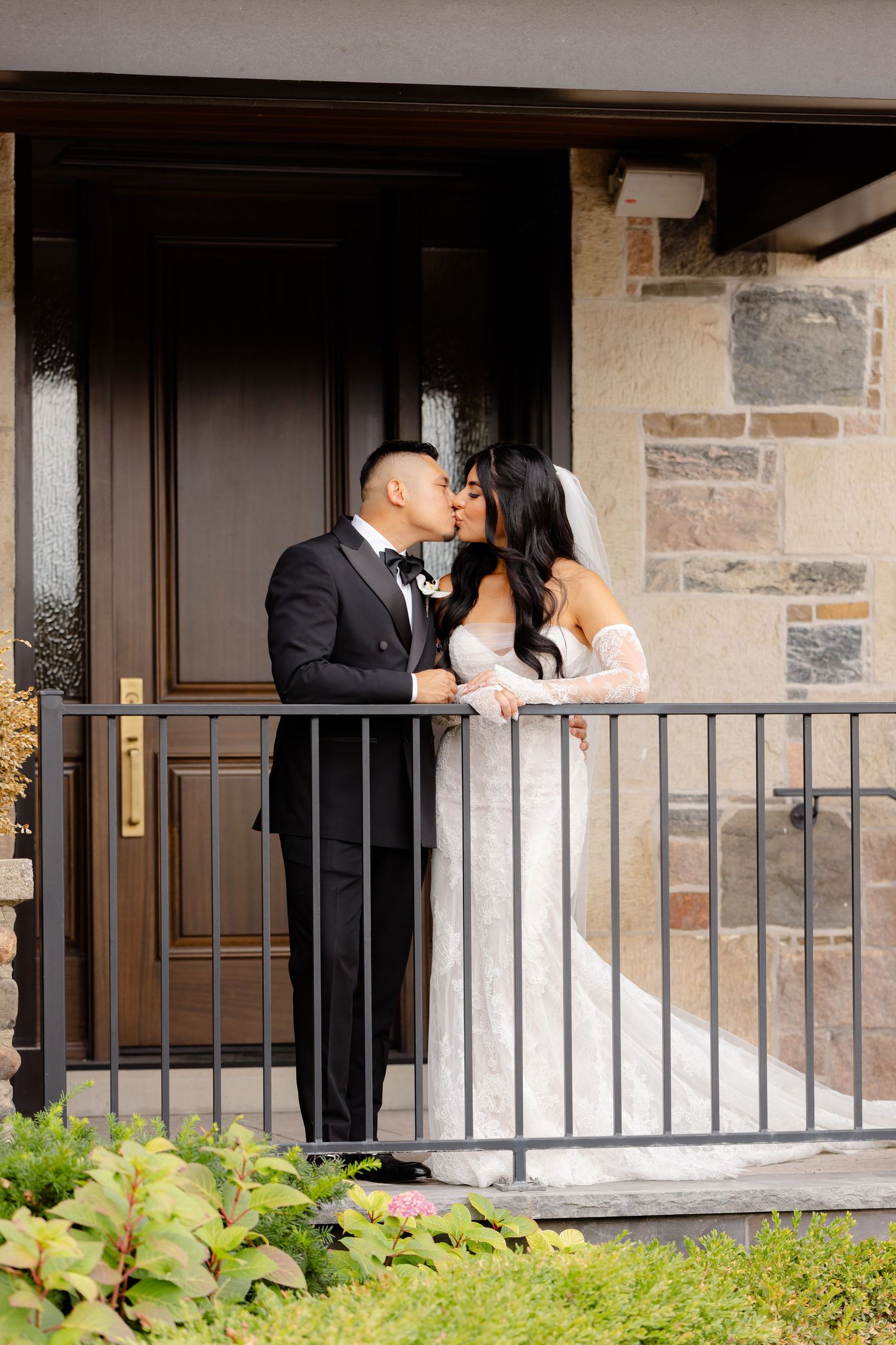 A romantic wedding moment captured as a couple shares a kiss on a stone porch with lush greenery in the foreground.