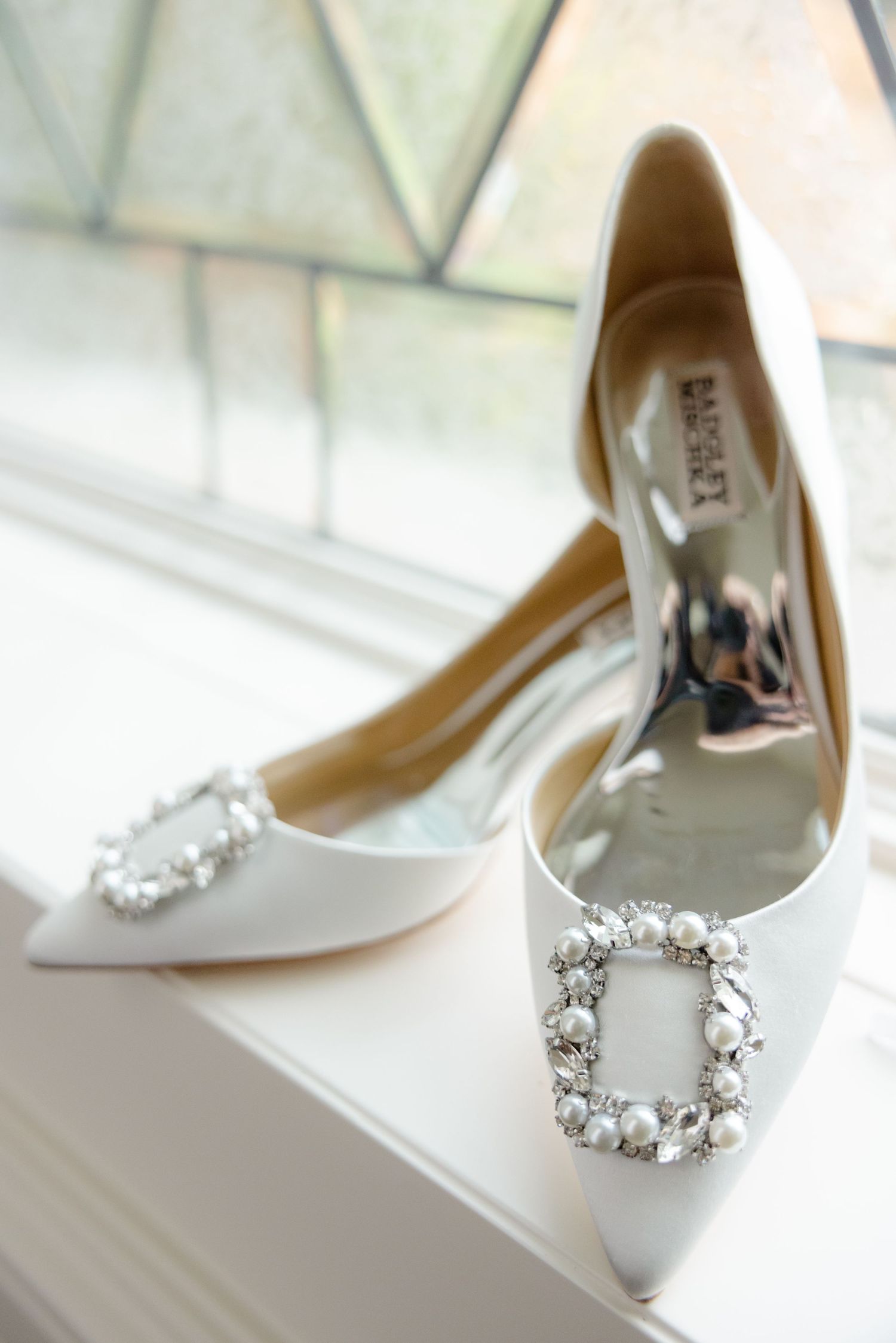 Elegant white bridal shoes with crystal embellished buckles displayed by a window.
