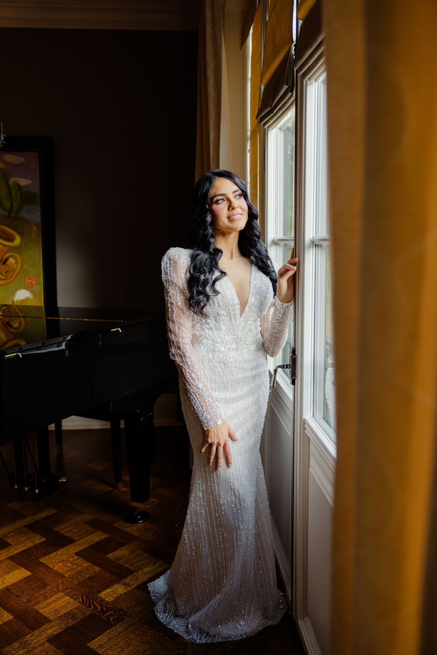 A reflective moment captured of a bride in a fitted lace gown standing by a sunny window.