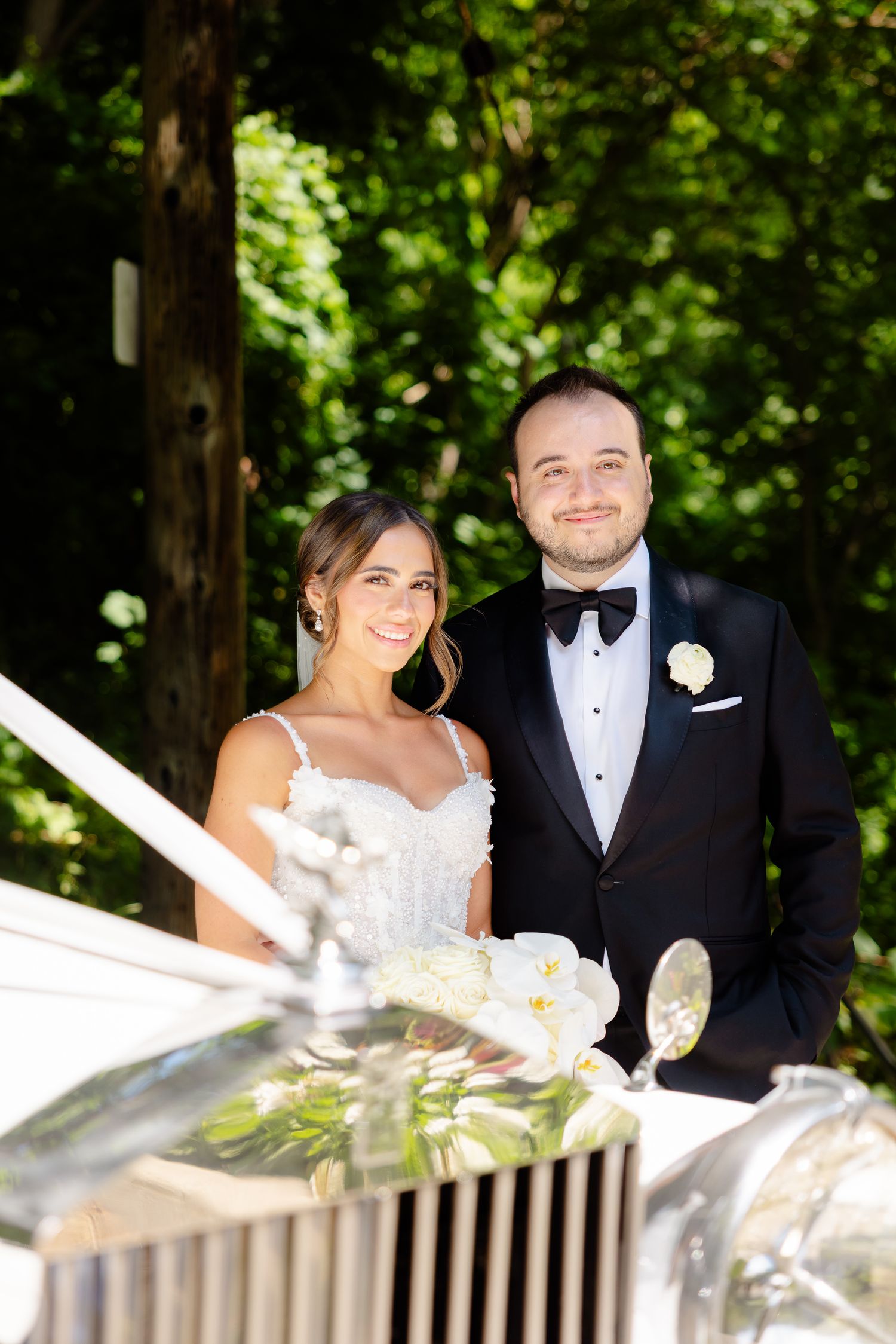 A happy elegant couple poses for a wedding photo next to a classic Rolls Royce car in a sunny outdoor setting.