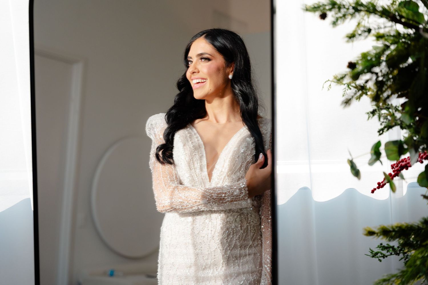 Smiling bride poses in white lace gown with plunging neckline near sunlit window.