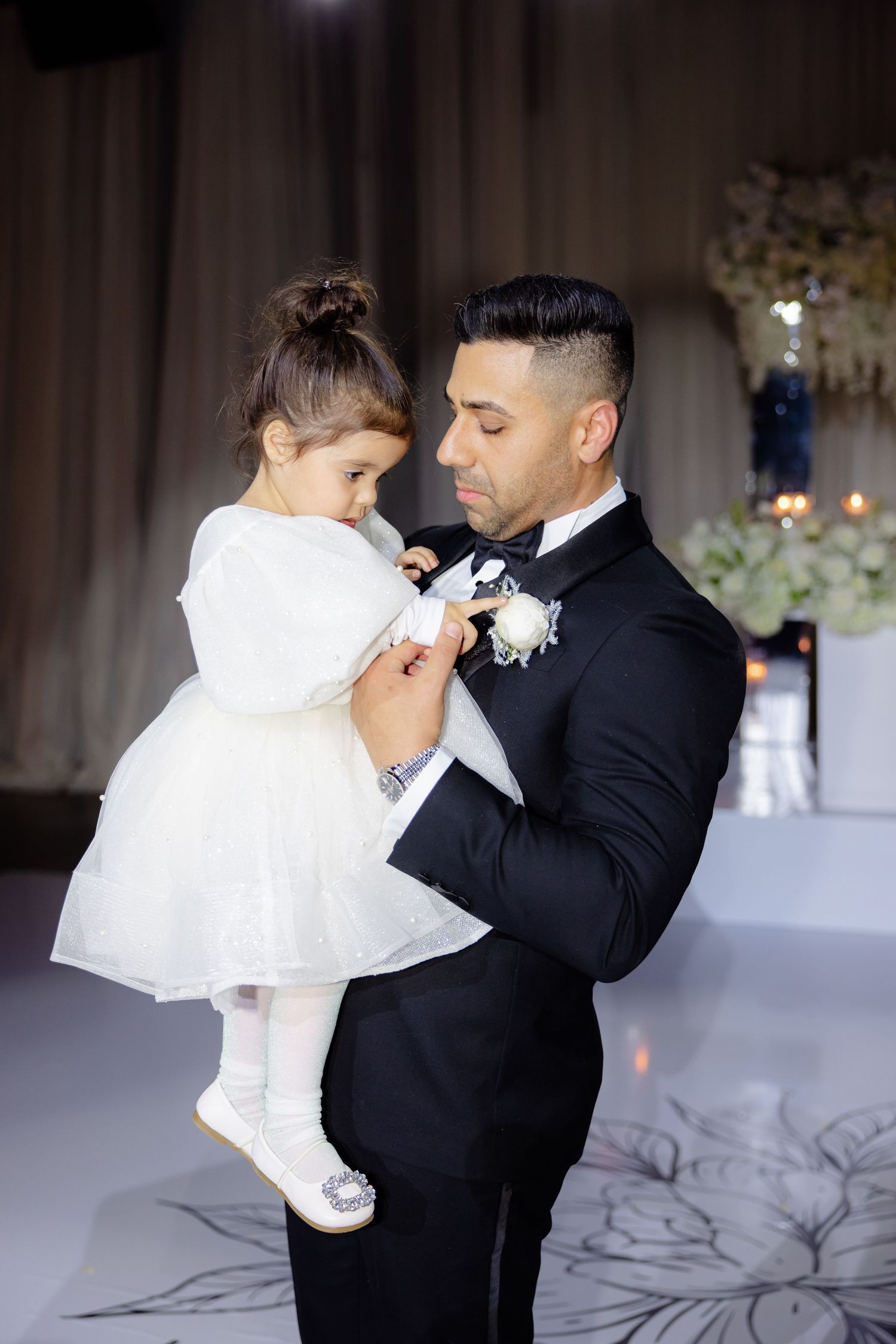 Person in tuxedo holds small child wearing white dress and cardigan at formal event.