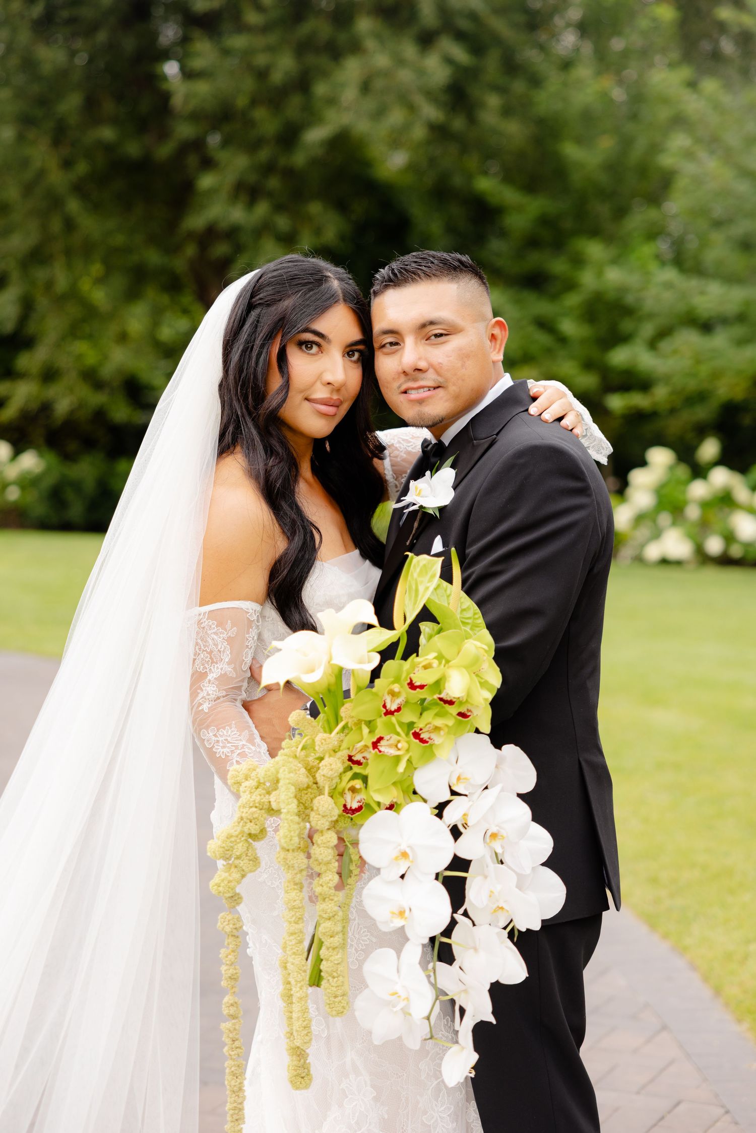 Romantic wedding couple poses together outdoors in a garden setting with orchid bouquet and white veil details.