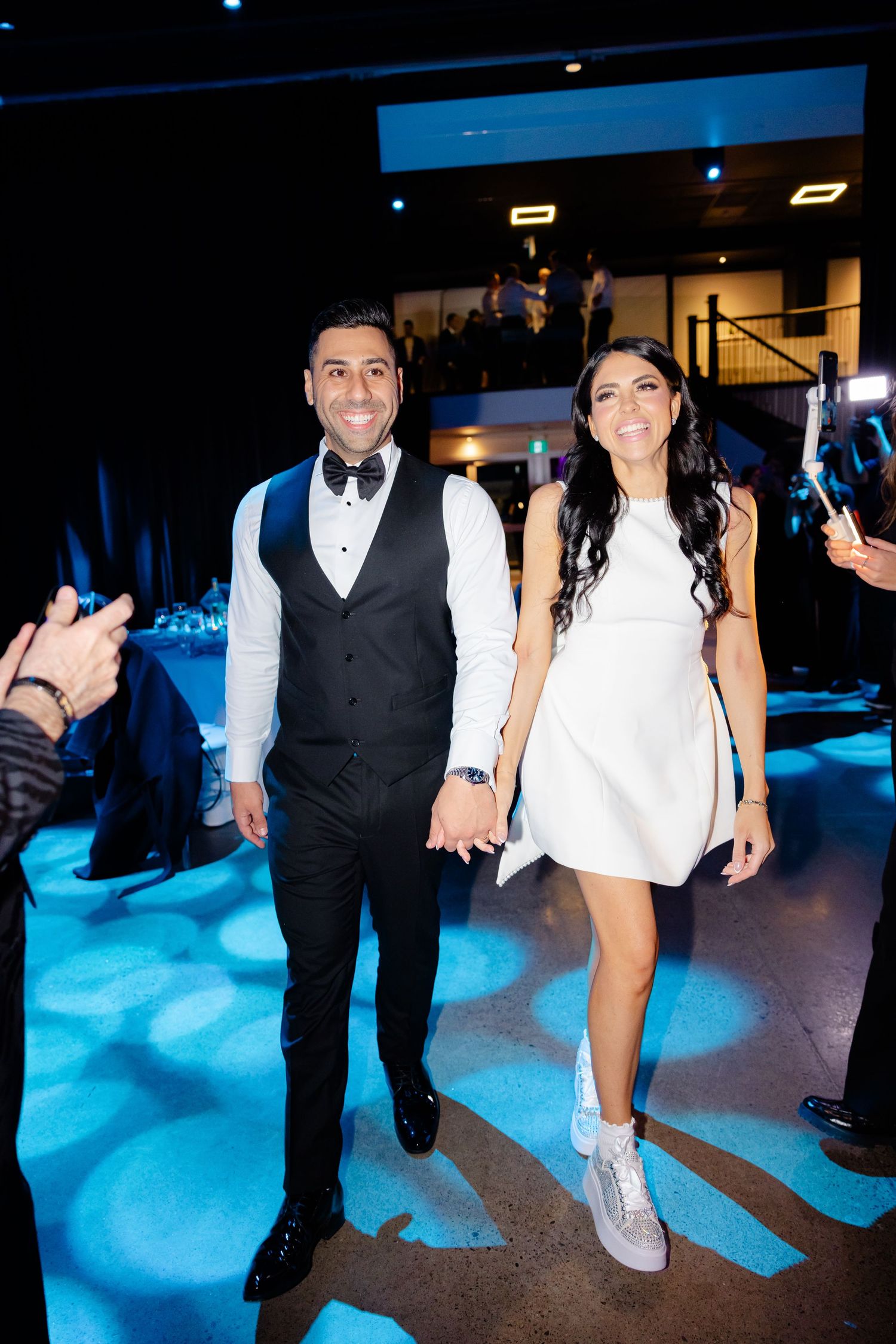 Wedding couple walks hand in hand across blue-lit reception floor.