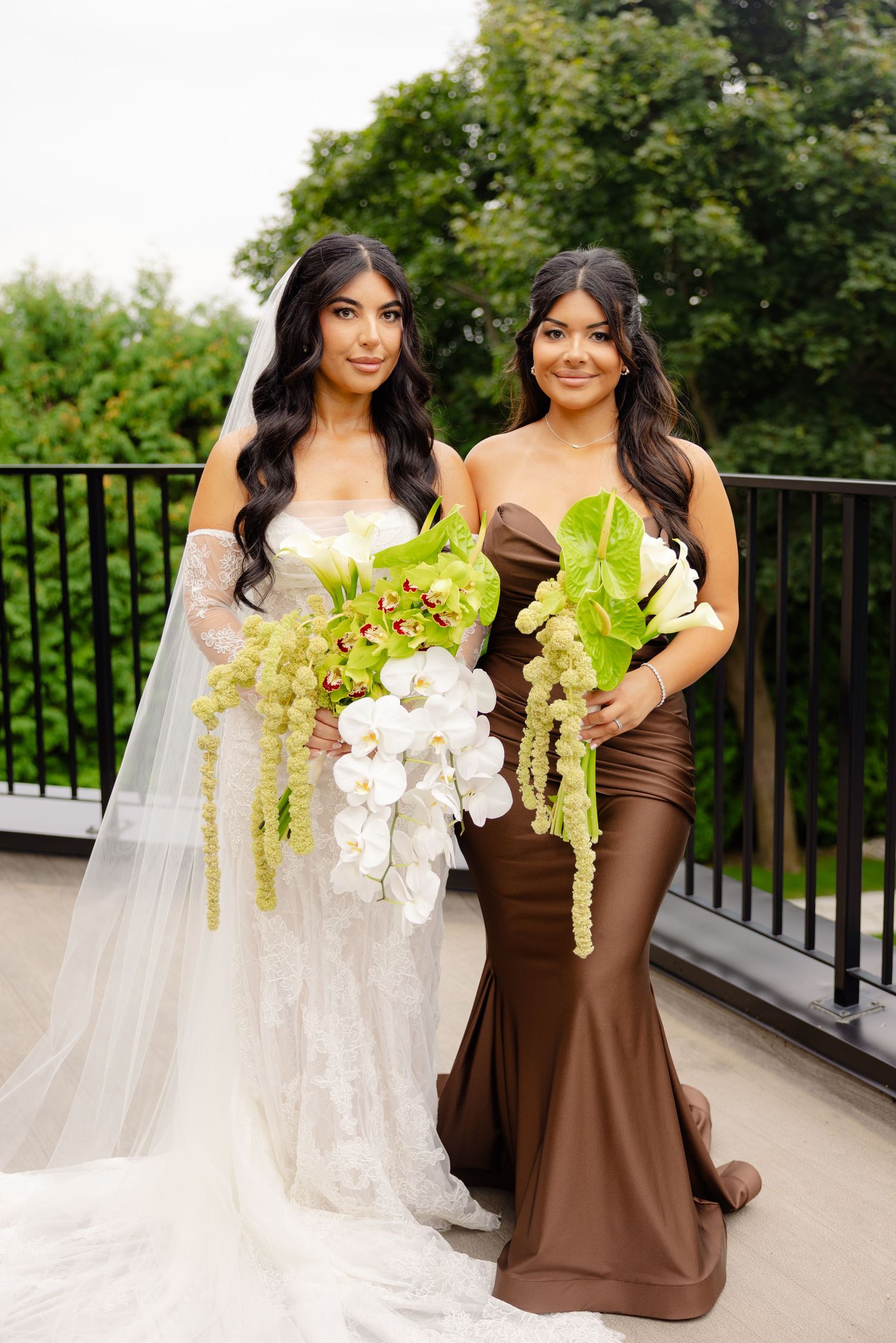 Bride in white lace gown and bridesmaid in brown satin dress hold stunning green and white floral bouquets on balcony.
